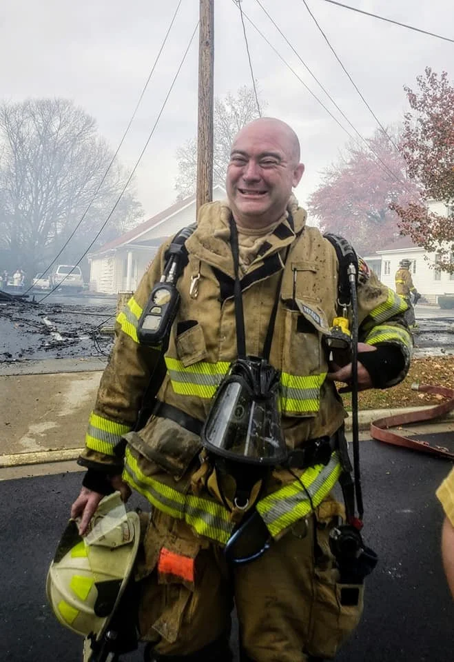 Smiling firefighter in firefighting gear stands in front of a house fire scene with smoke and charred debris in the background.