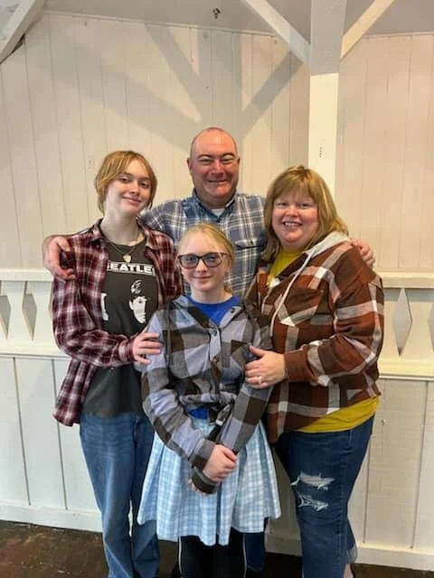 A group of four people, three girls and one man, standing close together indoors with a white wooden wall behind them. They are smiling and posing for the photo.
