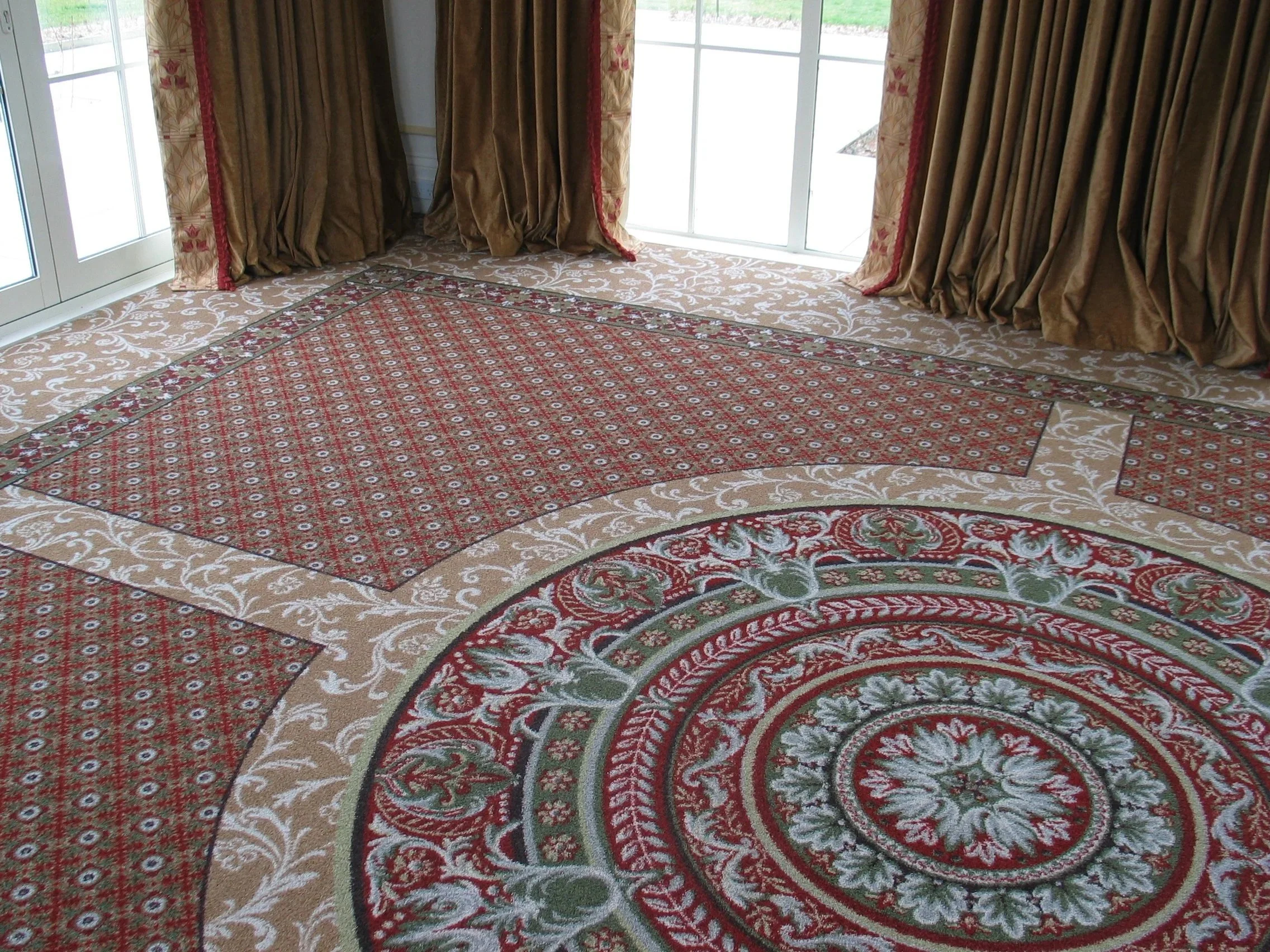 Elegant living room with a large ornate red and green patterned area rug on the carpeted floor, beige curtains with red and gold accents, and glass sliding doors letting in natural light.