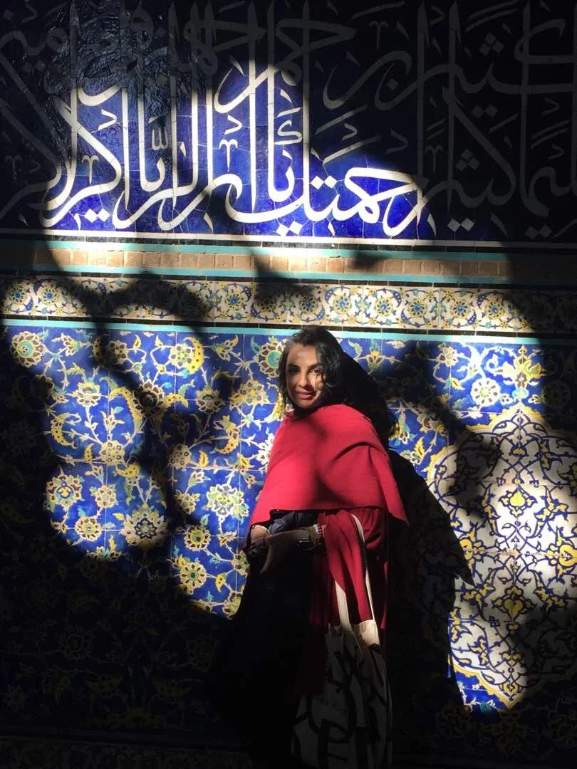 A woman in a red top and dark pants standing in front of a wall with blue and yellow patterned tiles, illuminated by sunlight and partially shaded by tree leaves. There is Arabic calligraphy on the wall.