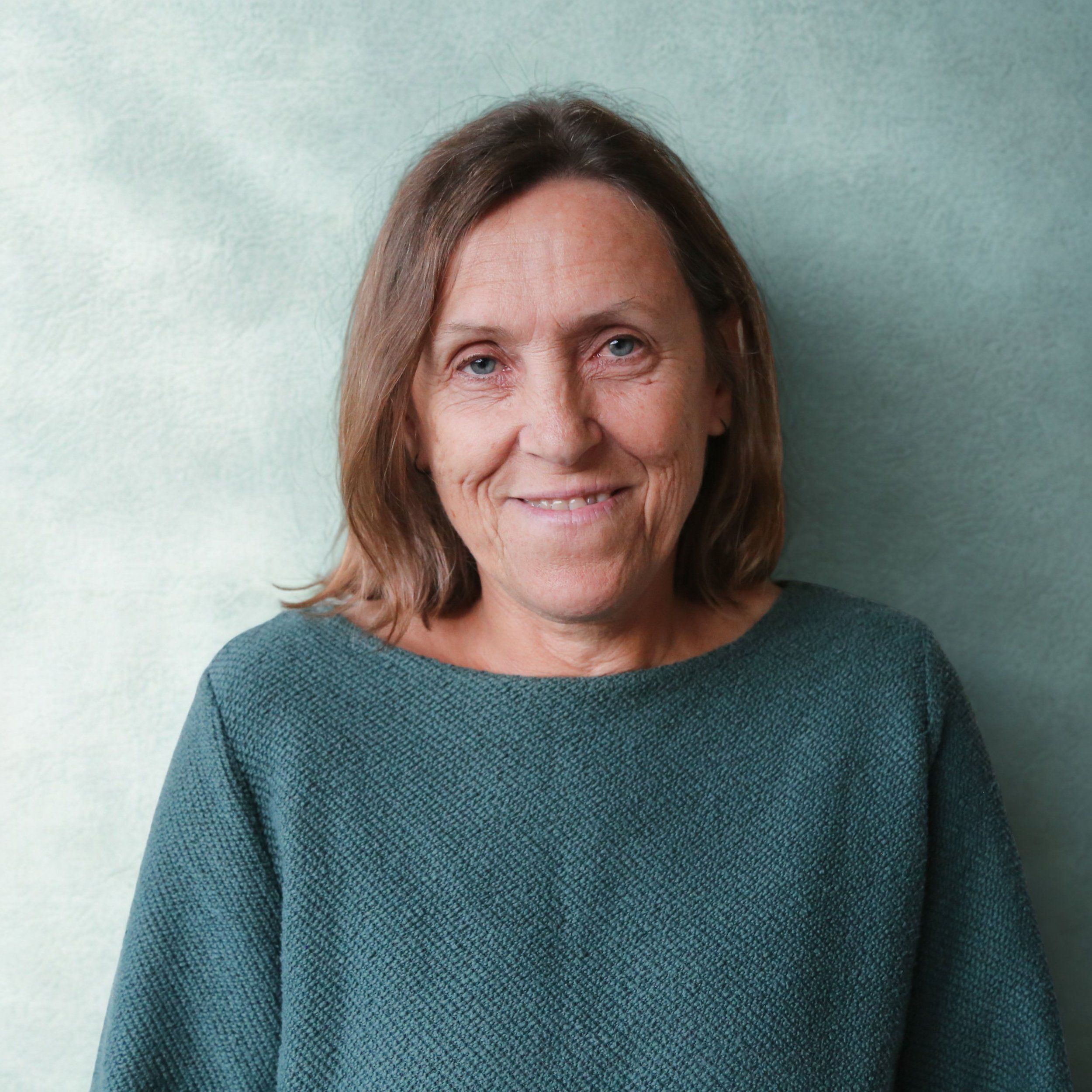 Partial view of a woman with light blue eyes, light brown hair, and fair skin, wearing a dark green textured sweater, standing against a light blue-green wall.