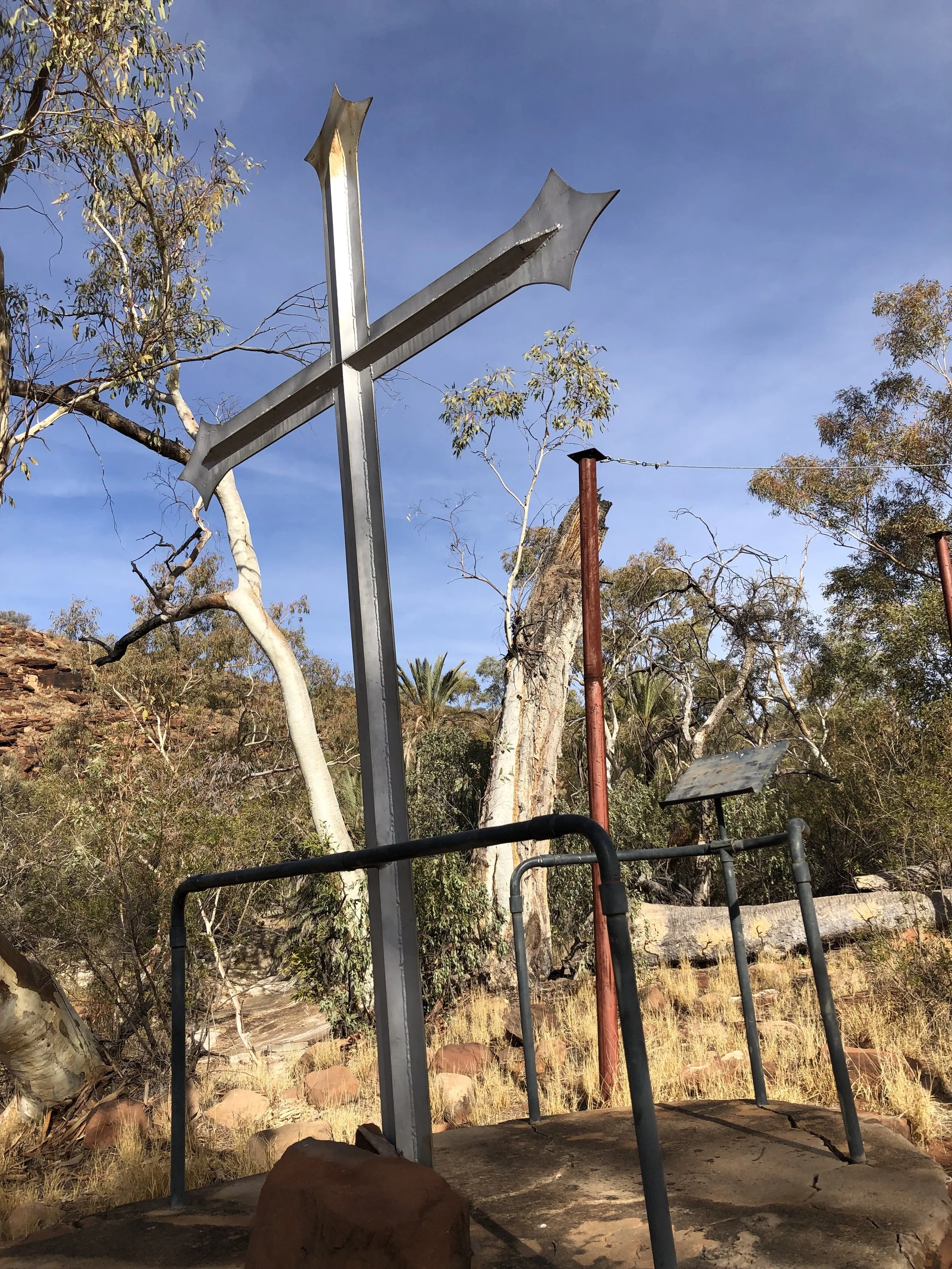 An outdoor scene with a metal cross-shaped sculpture, trees, dried grasses, and rocks, under a clear blue sky.
