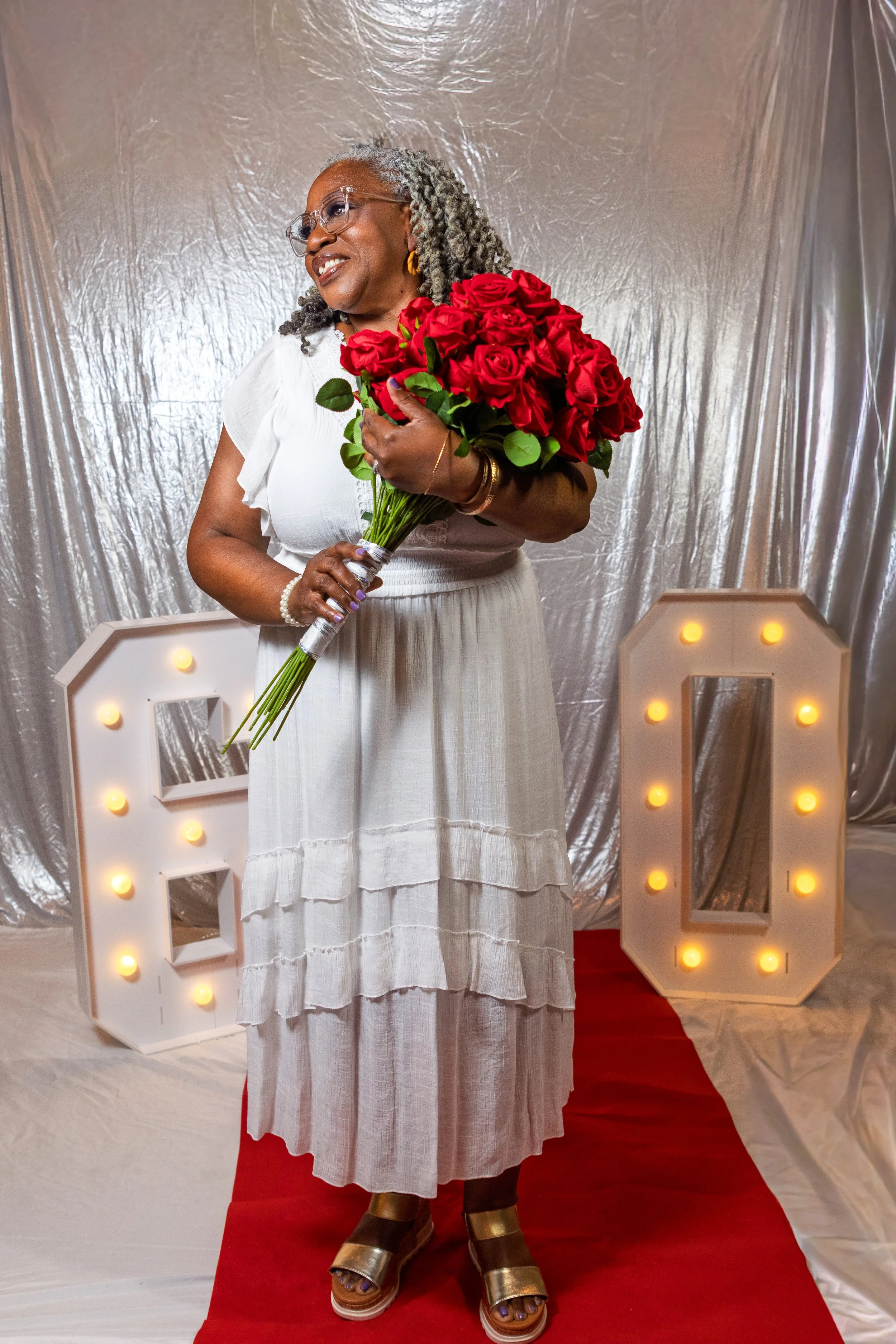 An older woman with gray curly hair, glasses, and gold earrings, dressed in a white dress, holding a bouquet of red roses, standing on a red carpet in front of illuminated marquee letters.