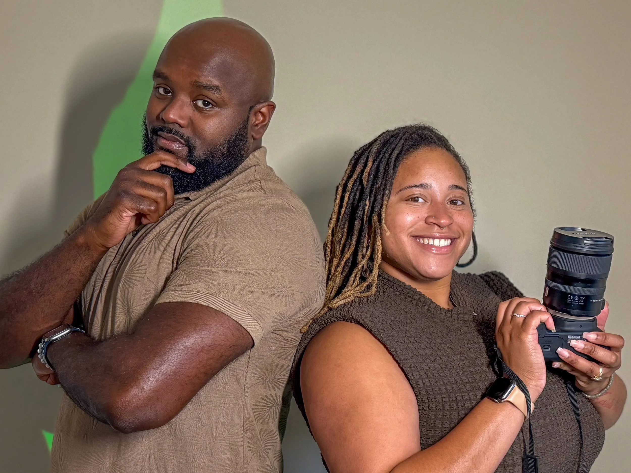 A man and a woman stand back-to-back, both smiling and looking at the camera. The man has a beard and is holding his chin with one hand, while the woman has dreadlocks and is holding a camera lens.