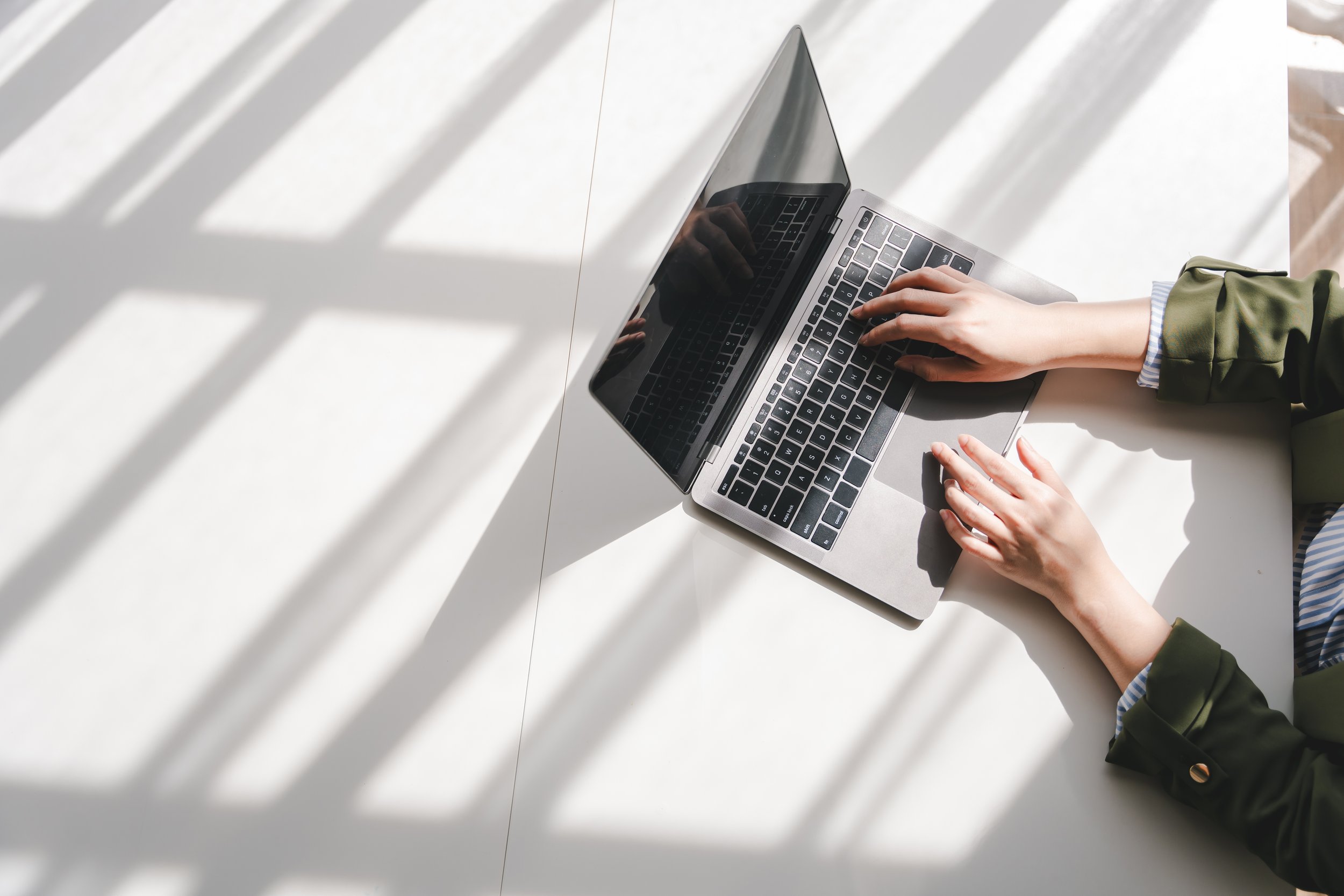 Person working on a laptop at a white desk with sunlight and shadows creating parallel lines across the surface.