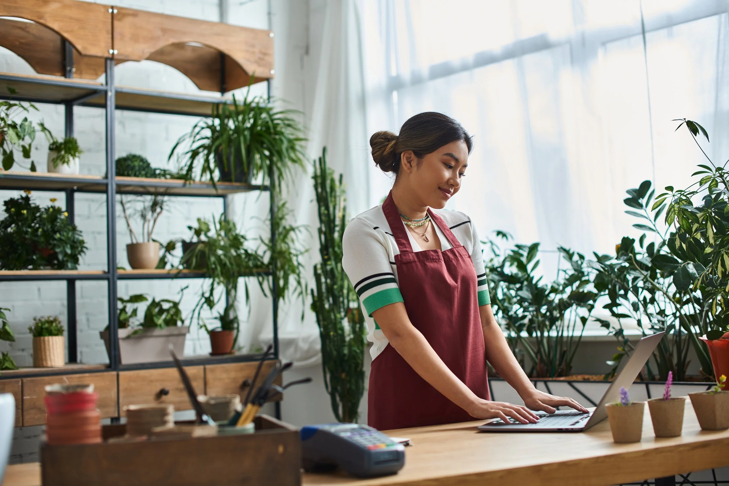 A woman in a red apron working with plants and a laptop in a bright greenhouse or plant shop with shelves of potted plants.