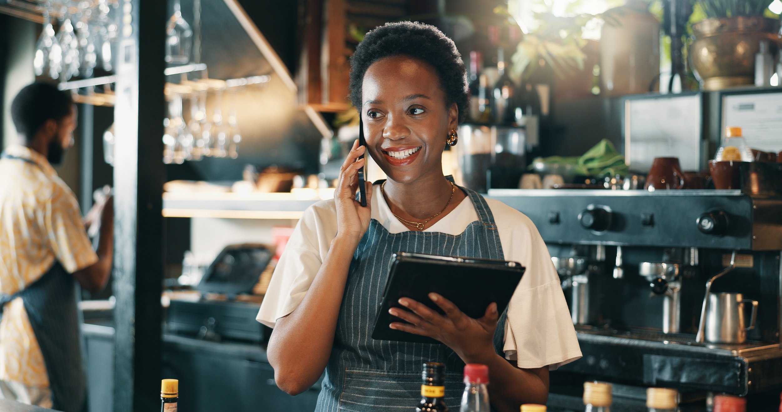 A smiling woman in a striped apron talking on her phone while holding a notepad in a busy cafe kitchen.
