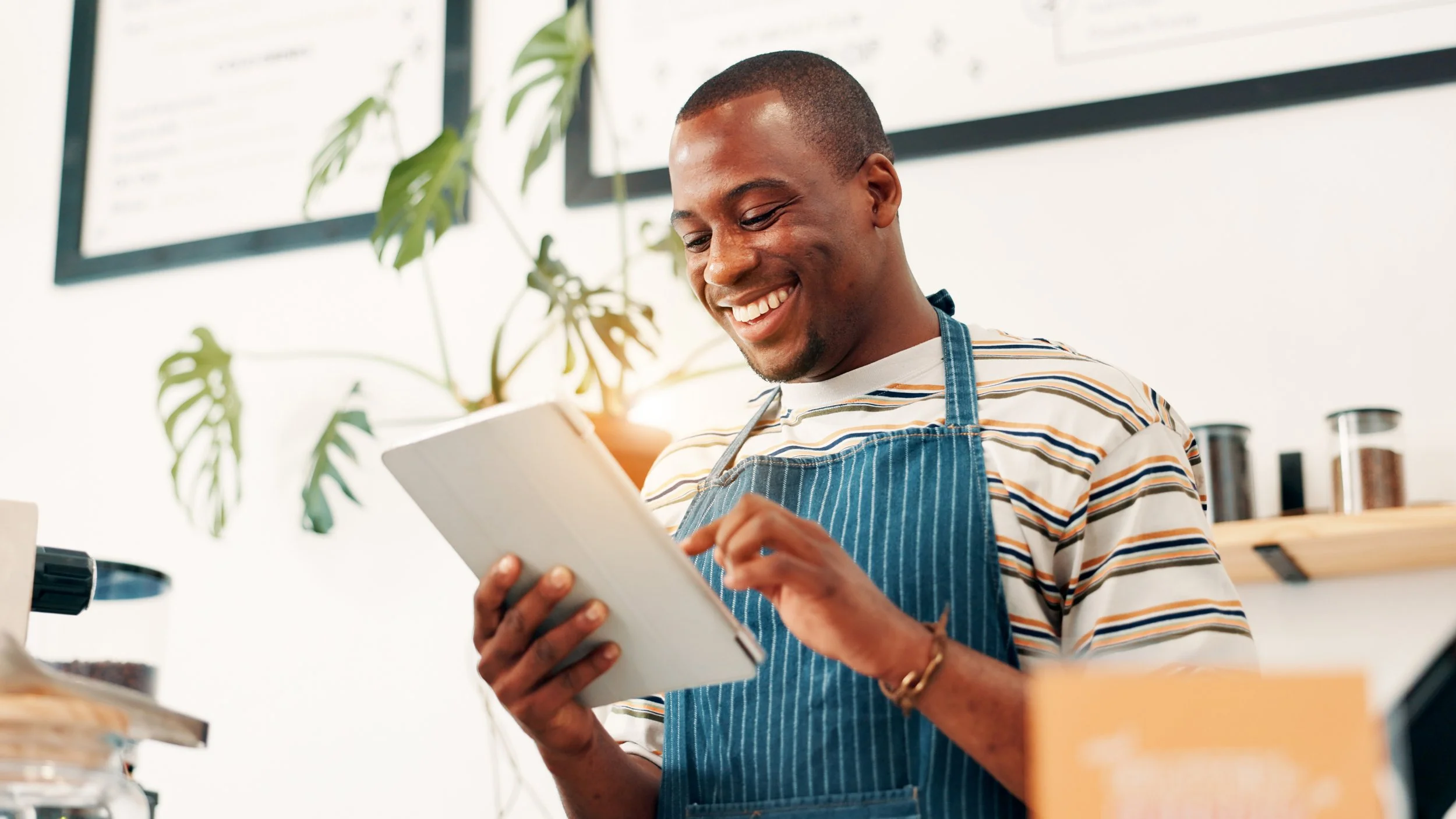 A smiling man in a striped shirt and denim apron looking at a tablet in a bright, modern workspace with plants and jars in the background.