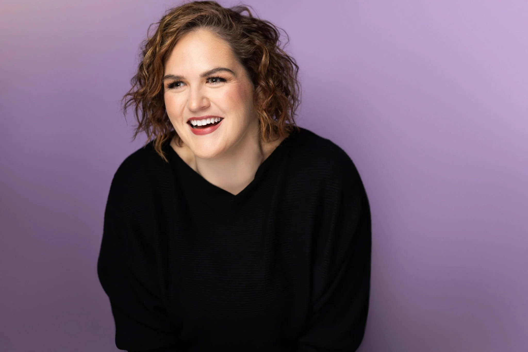 A woman with curly, shoulder-length, reddish-brown hair smiling and looking slightly to her right against a purple background, wearing a black top.