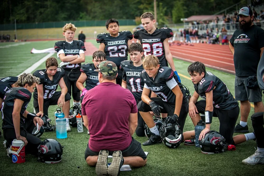 Football coach kneels on the field talking to a youth football team after a game.