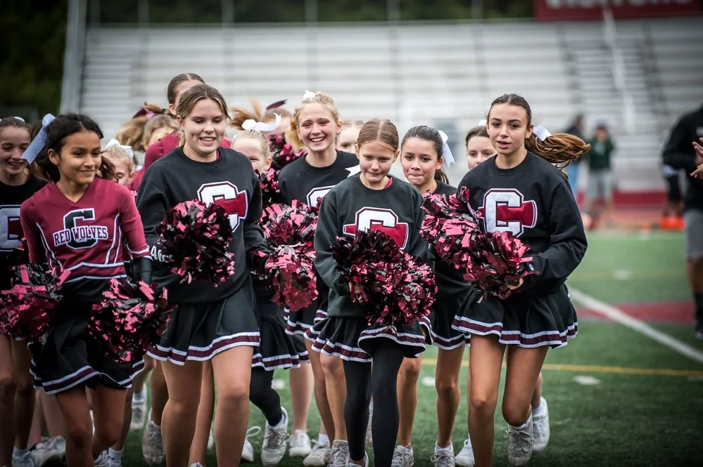Group of cheerleaders in black and crimson uniforms smiling and holding pom-poms on a football field