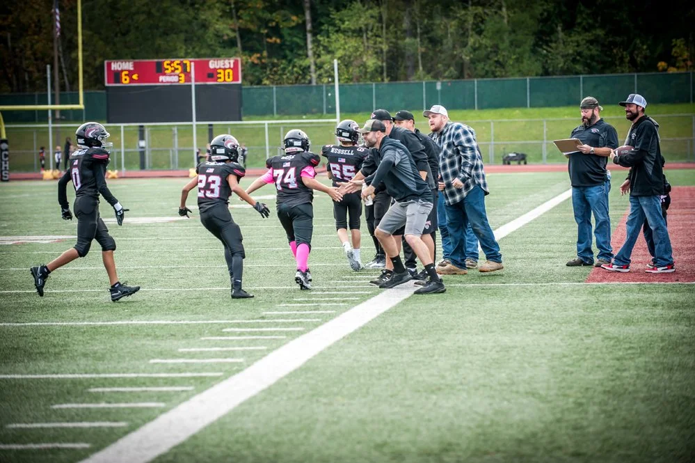 Youth football players in black uniforms celebrate with coaches on a football field.
