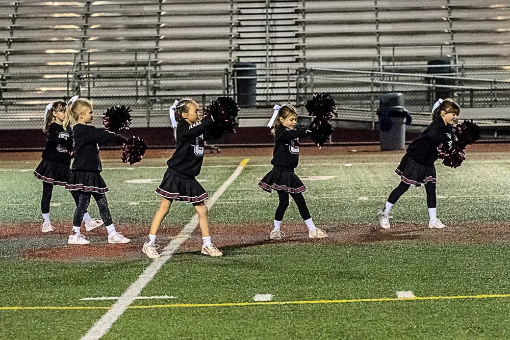 Girls cheerleaders performing on a football field at night, holding pom-poms and wearing matching black outfits.