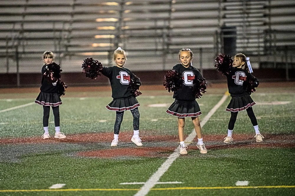 Four young cheerleaders in black uniforms with red and white accents, holding pom-poms, standing on a football field at night.