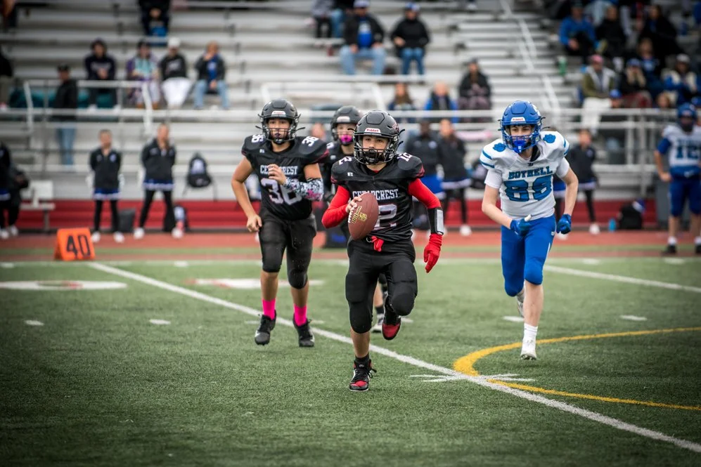 Young football players in action on the field, with one carrying the ball and others running behind, during a game at a stadium with spectators in the background.