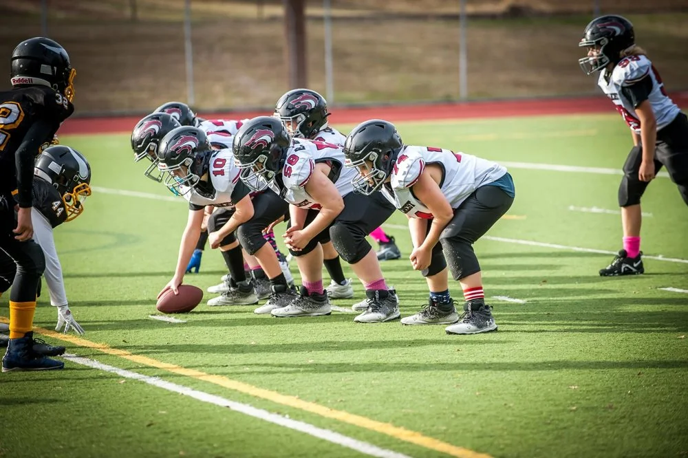 Young football players in helmets and gear line up in formation on a football field, ready for play.