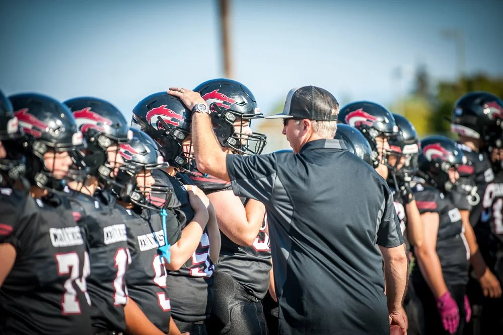 A football coach giving a pep talk to a team of young players in black uniforms with helmets during daytime.