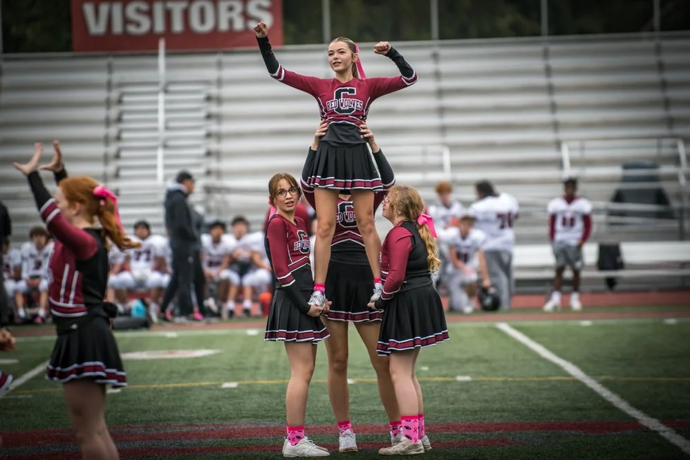 Cheerleaders performing a routine on a football field with spectators in the bleachers in the background.