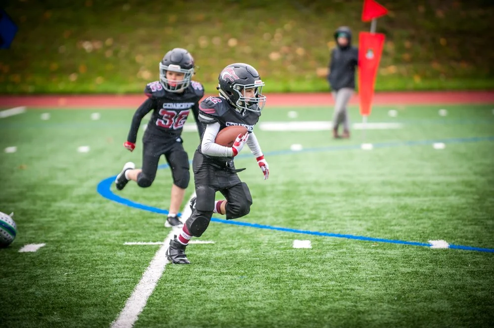 Young children in football gear playing on a football field during daytime, with one child running with the ball and others pursuing.