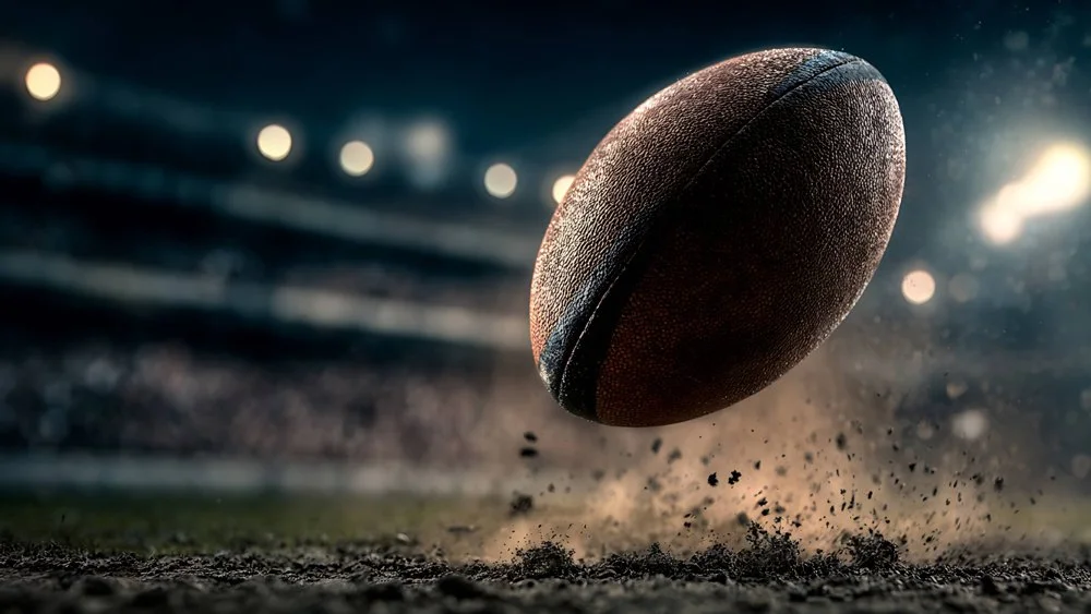 A brown leather football bouncing on a dirt field under stadium lights at night.