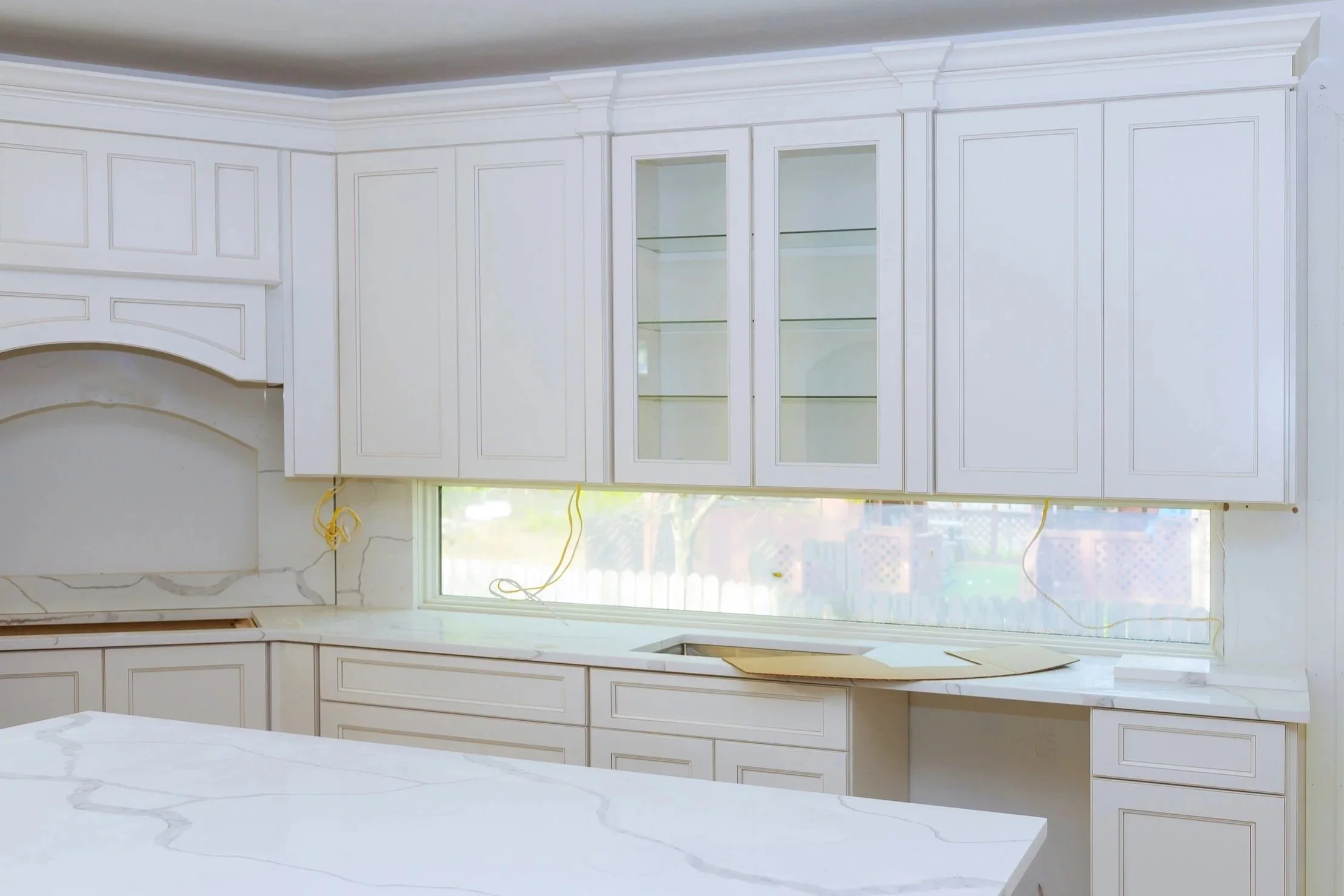 A kitchen under renovation with white cabinetry and marble countertops, no appliances installed yet, with exposed yellow electrical wiring and a large window overlooking a backyard.