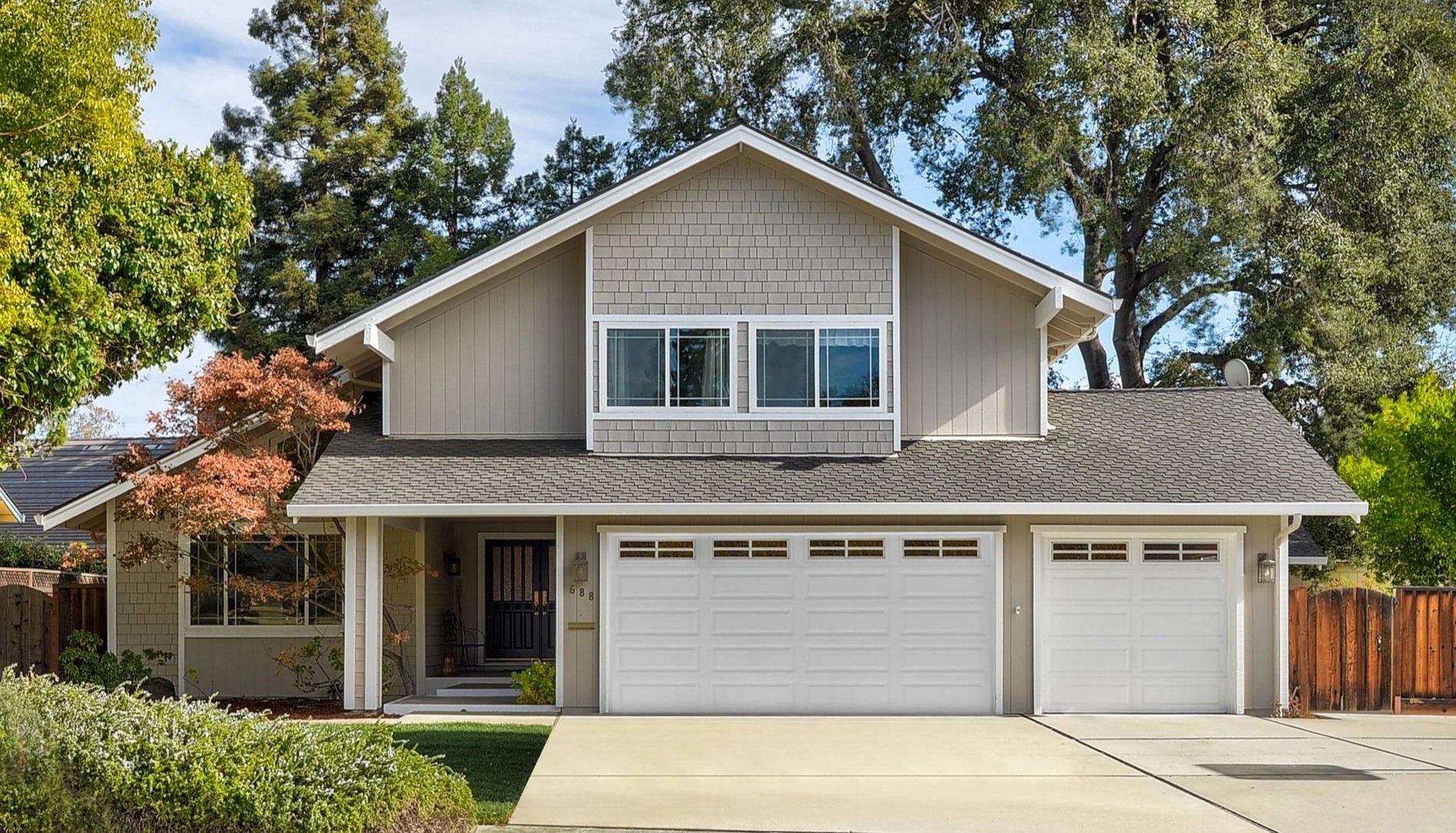 A two-story suburban house with a grey shingle roof, white garage doors, and a black front door. There are trees and bushes surrounding the house, and the driveway is concrete.