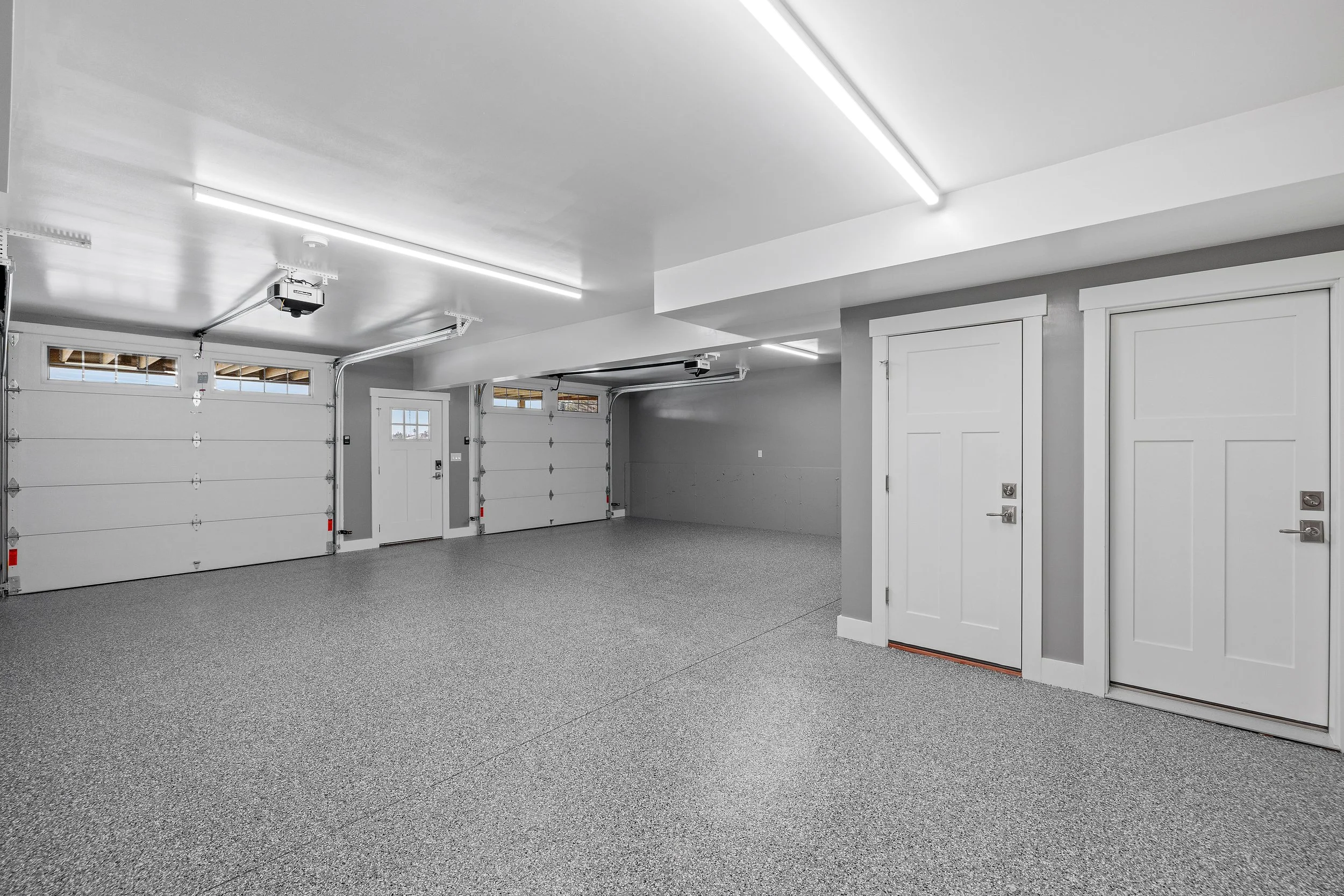 Empty clean garage with speckled gray epoxy floors, white panel garage doors, small windows, and bright ceiling lighting.