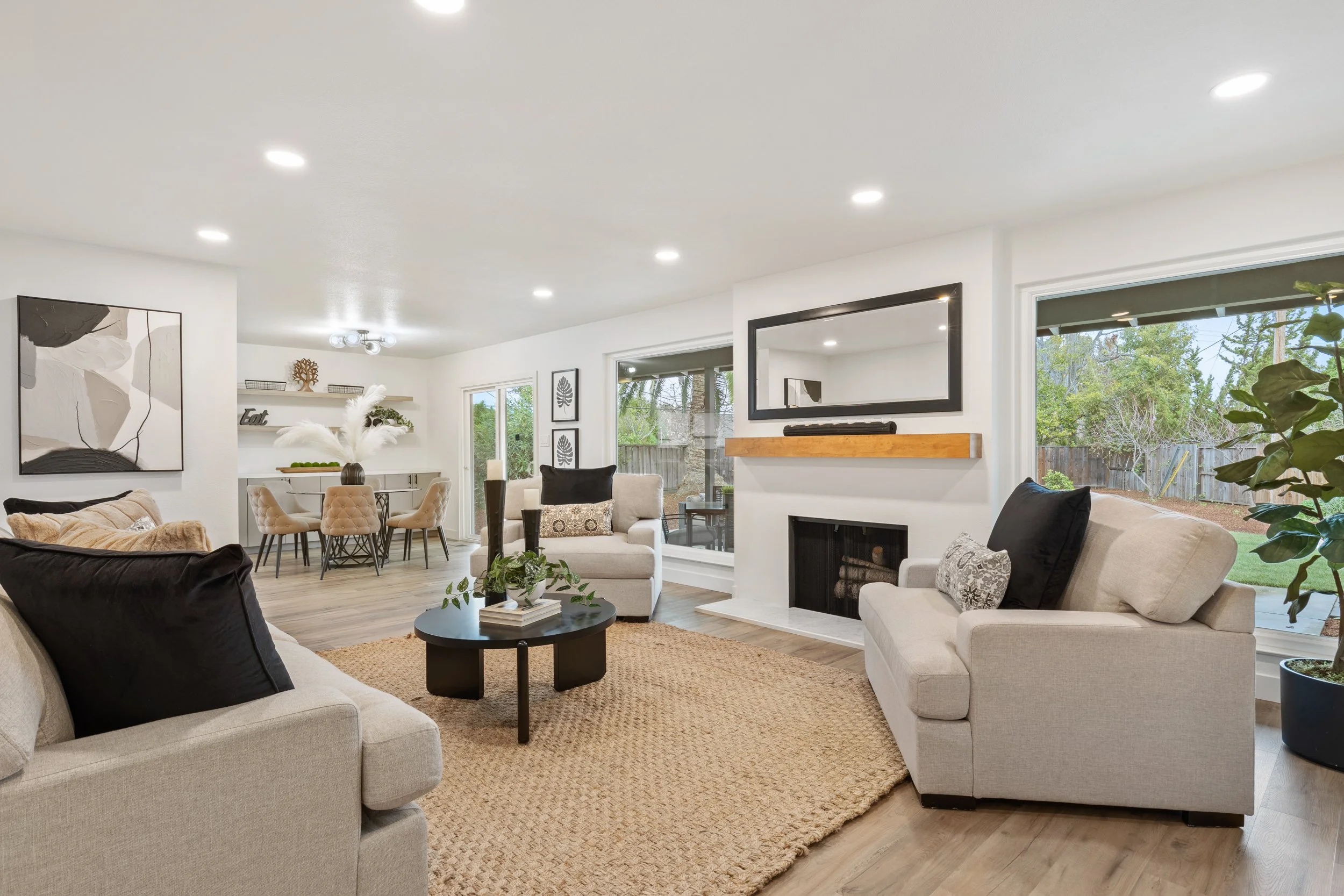 Bright, modern living room with beige sofas, black pillows, a black coffee table, a fireplace, large windows showing a backyard, and minimal decor.