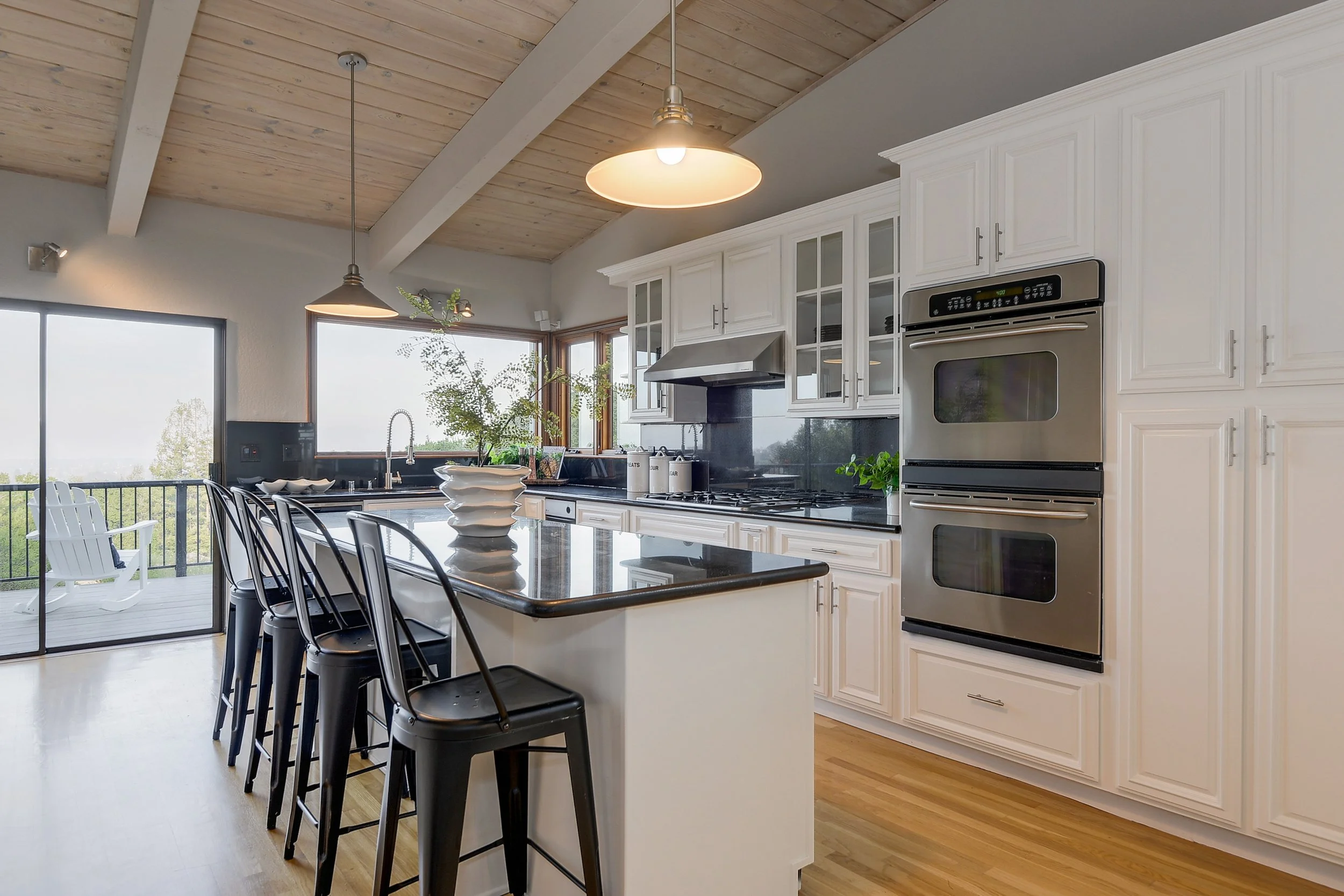 Modern kitchen with white cabinets, black countertop island, stainless steel double oven, gas stove, and large windows with a view of a deck and outdoor chairs.