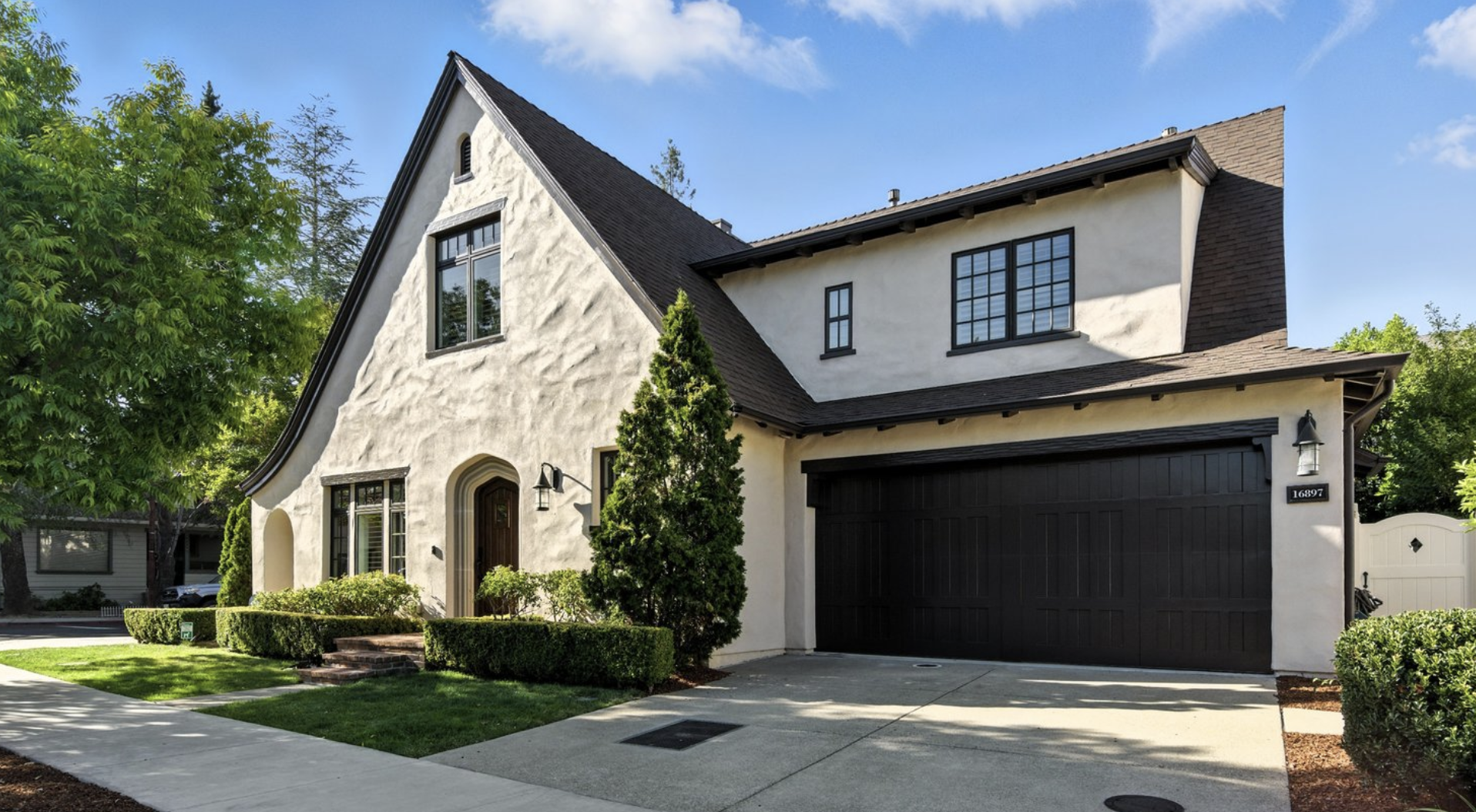 A two-story house with white stucco exterior and a dark brown shingled roof. It has a black garage door, a small front yard with greenery and trees, and a clear blue sky.