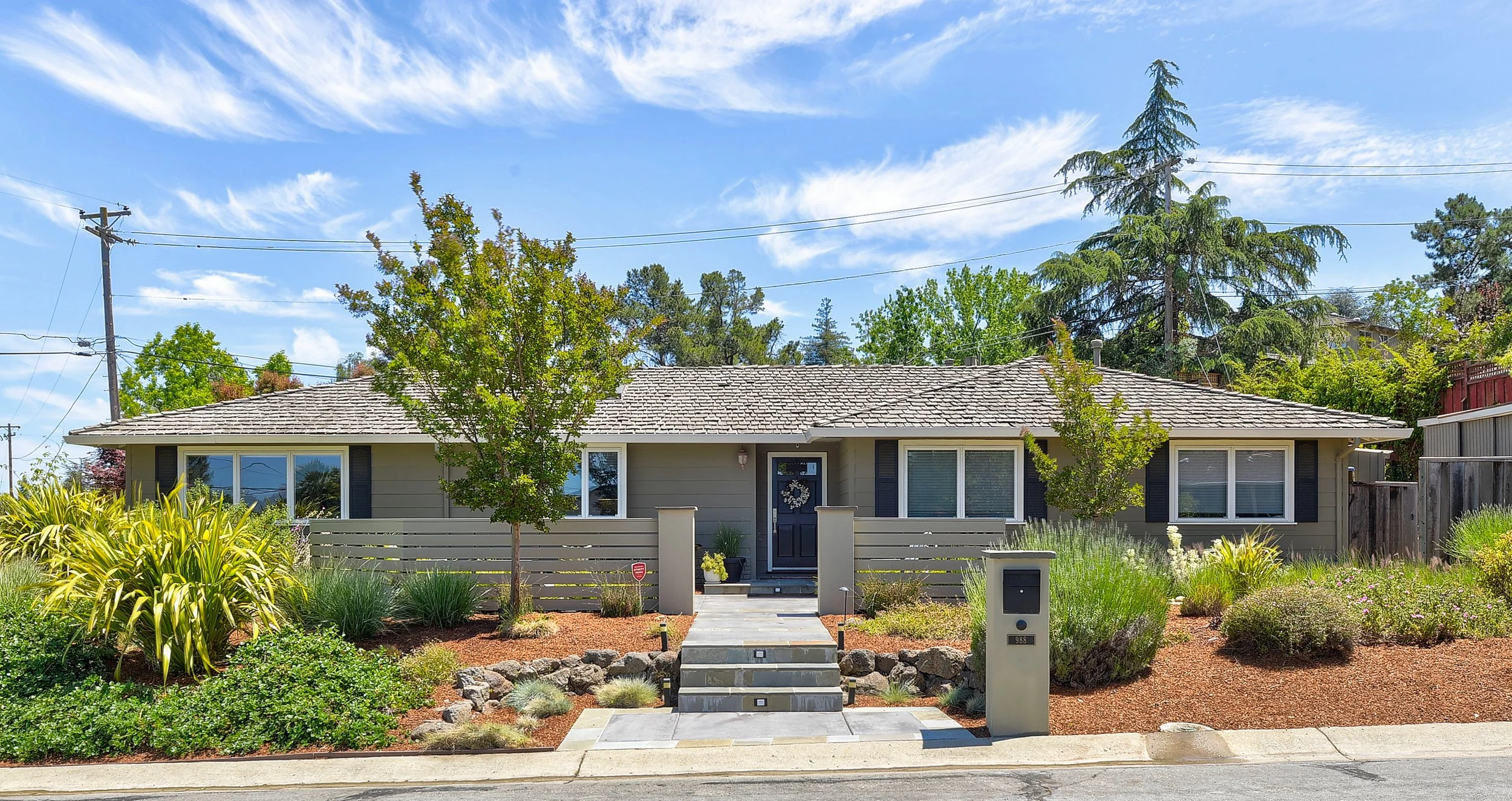 Single-story house with gray siding, black shutters, and a gray shingle roof. Front yard has trees, shrubs, and mulch, with a concrete walkway leading to a black front door with a wreath. Mailbox on sidewalk.