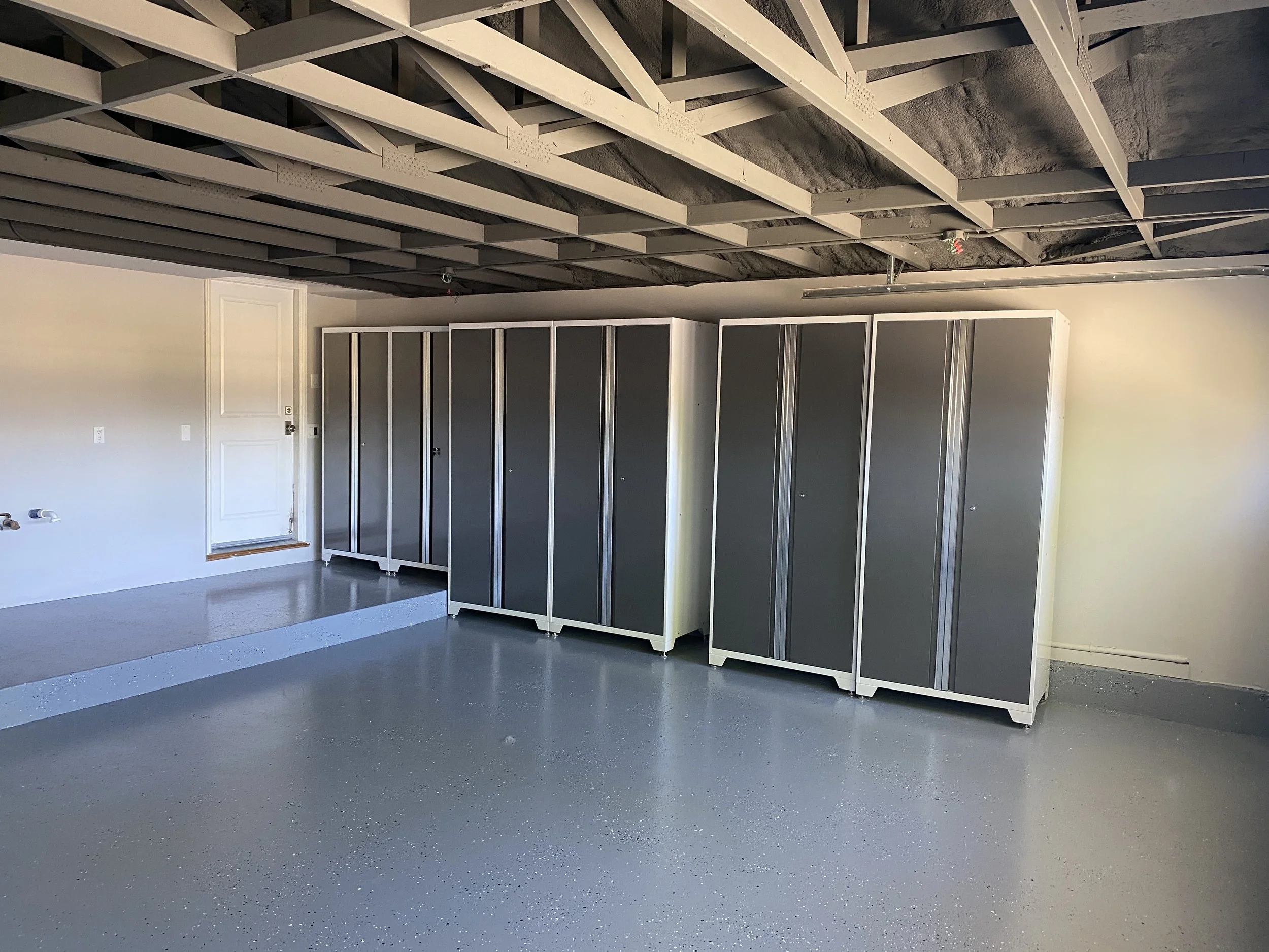 Empty garage with gray flooring, white walls, a white door, and multiple gray storage cabinets lined against the wall. The ceiling shows exposed beams and insulation.