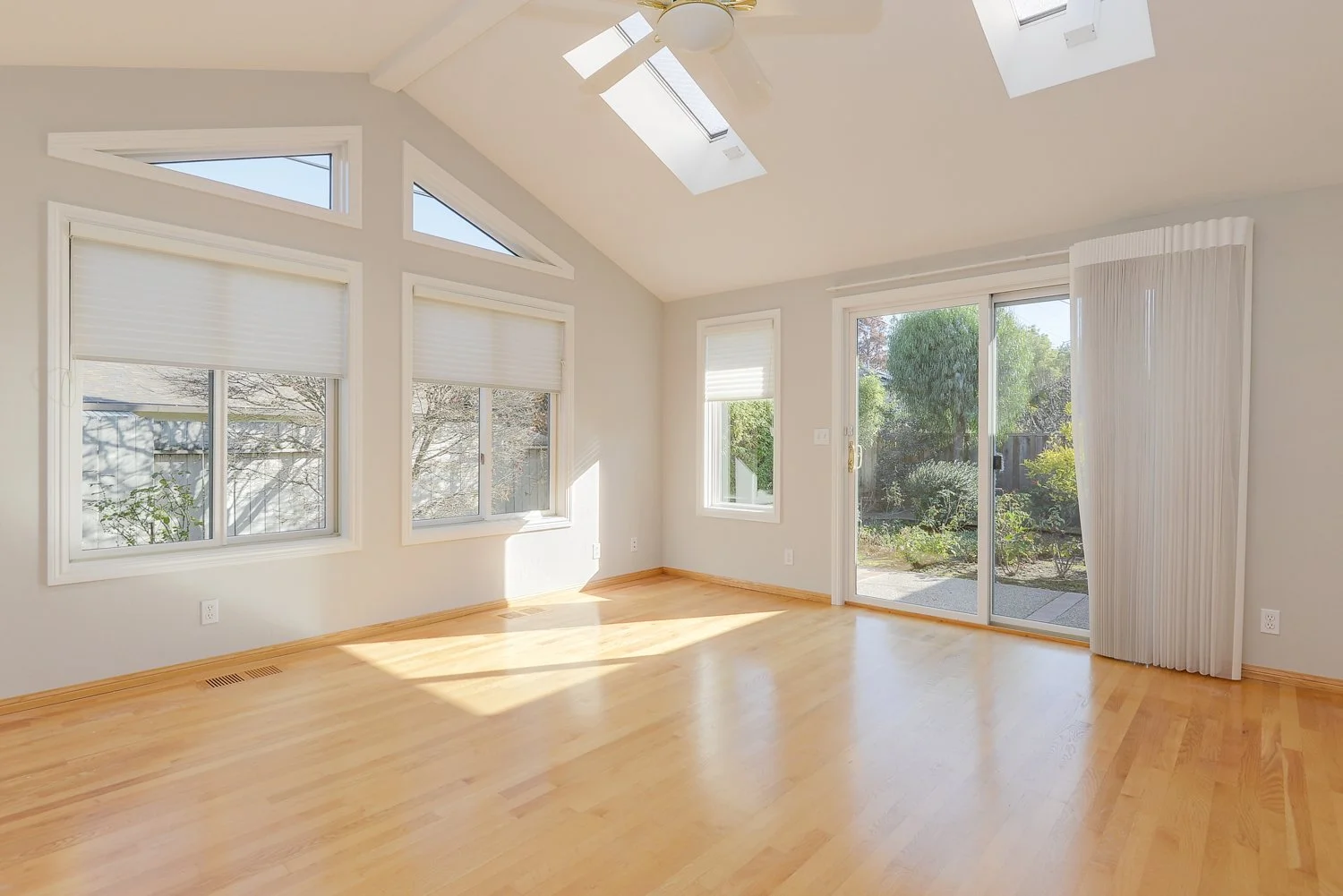 Empty living room with light wood floors, white walls, multiple windows with blinds, dotted with sunlight, a sliding glass door leading outside, and skylights in the ceiling.