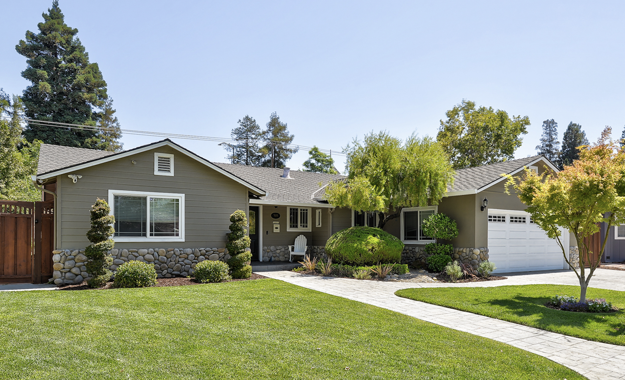 Front view of a suburban house with gray siding, stone accents, a white garage door, a walkway driveway, green lawn, and landscaped garden with trees and shrubs.
