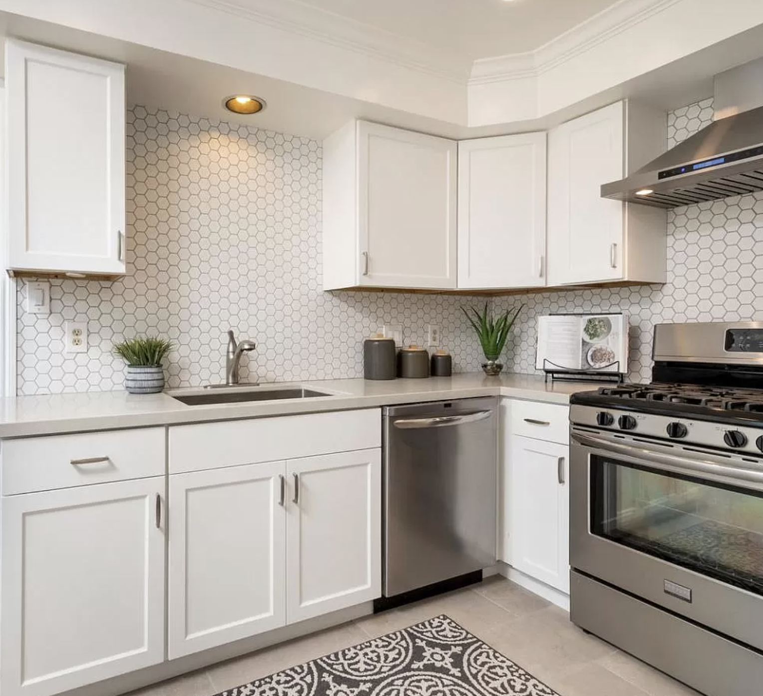 Modern kitchen with white cabinets, stainless steel dishwasher and oven, hexagon tile backsplash, small plants, and decorative jars on the countertop.