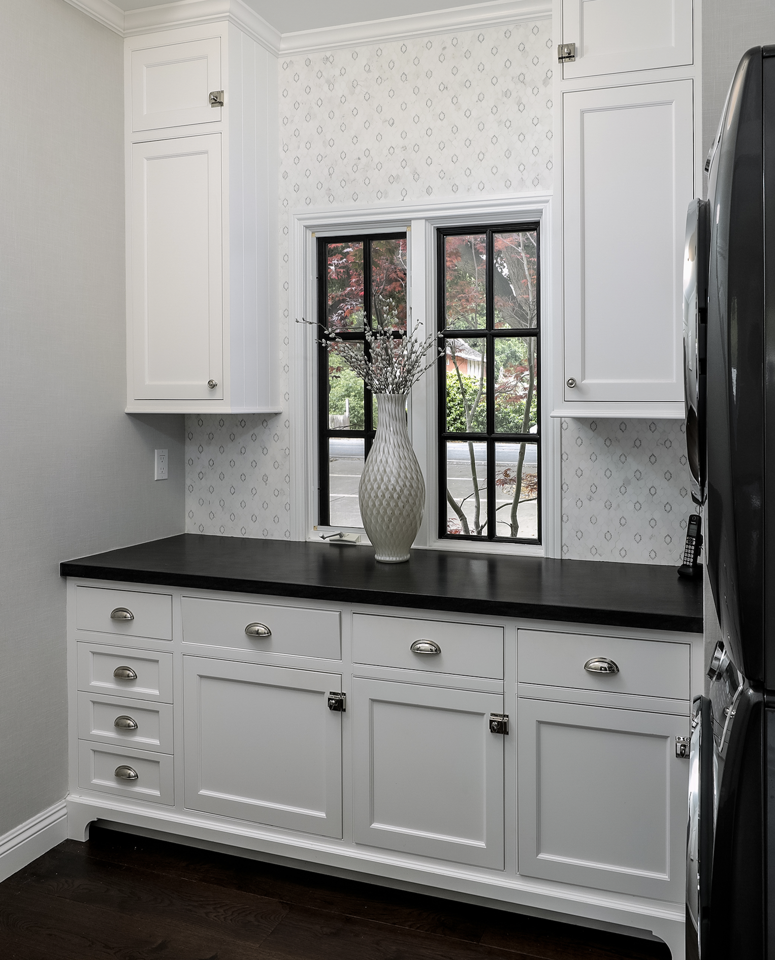 A laundry room with white cabinets, a black countertop, and a window with a vase of white flowers on it.