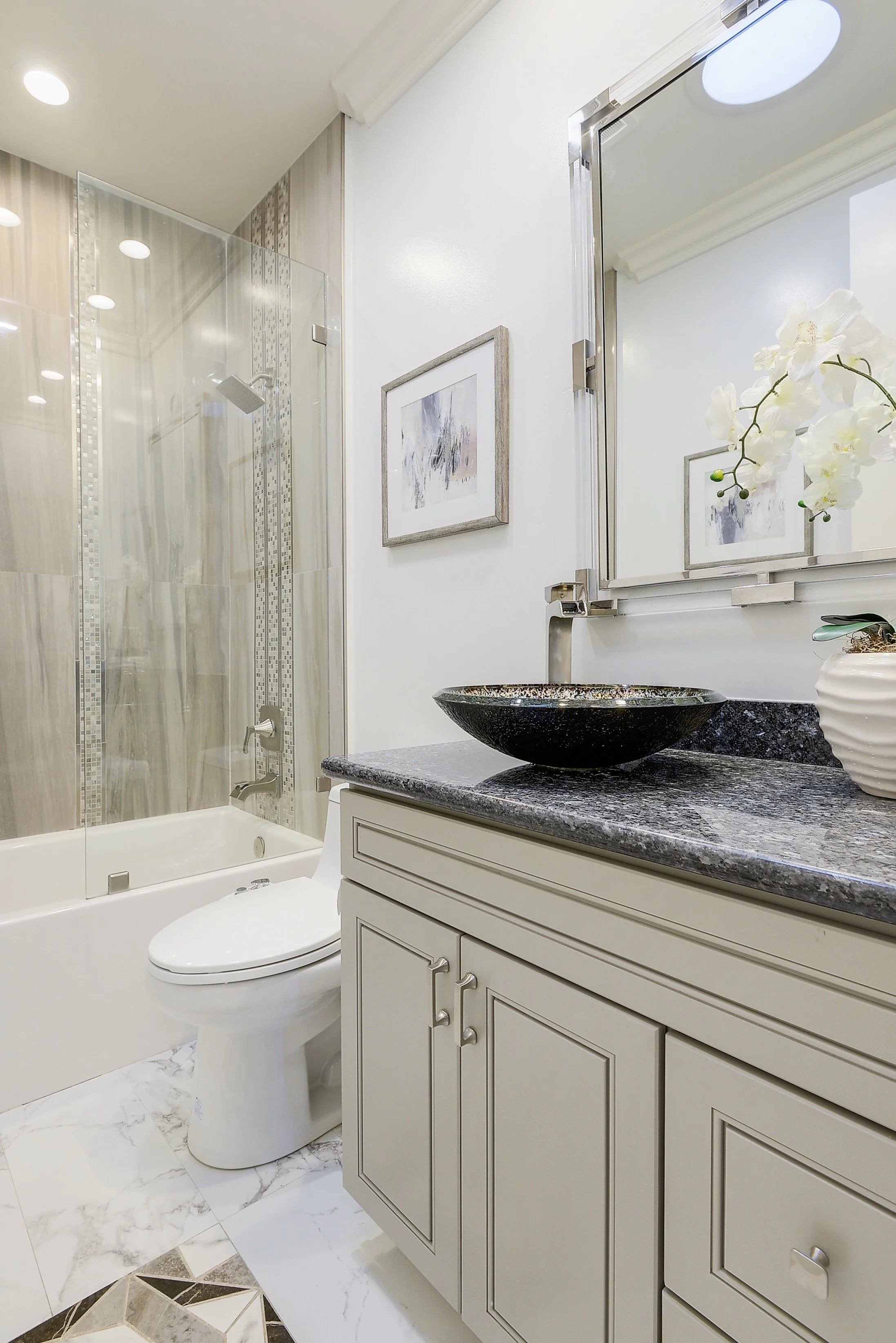 A modern bathroom with a white vanity cabinet, granite countertop, black vessel sink, and wall mirror. There is a marble tile floor, a framed picture on the wall, and a flower in a white vase. A shower with glass doors is in the background.