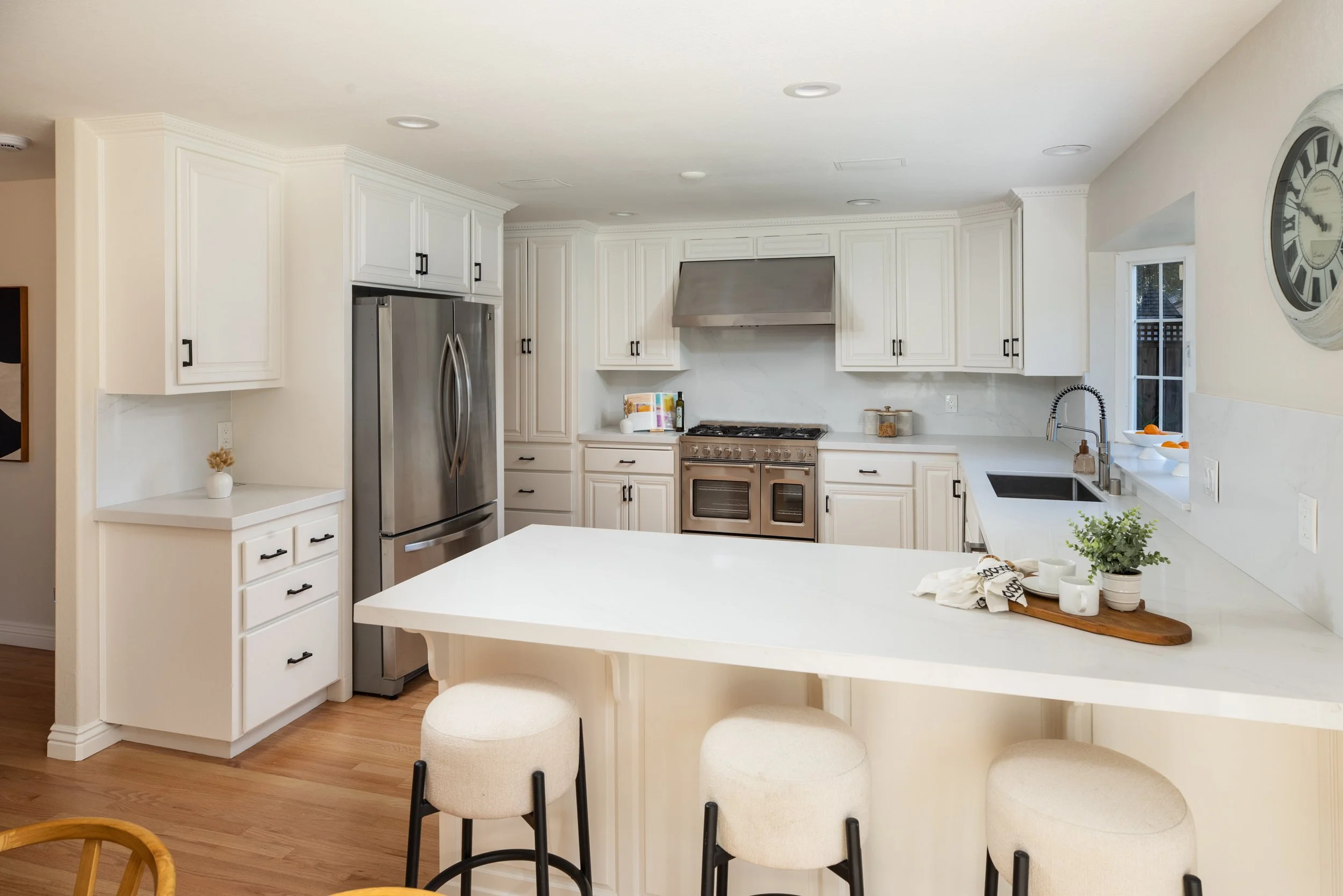 Modern white kitchen with stainless steel refrigerator, oven, and range hood, white cabinetry, window above sink, and a kitchen island with stools.
