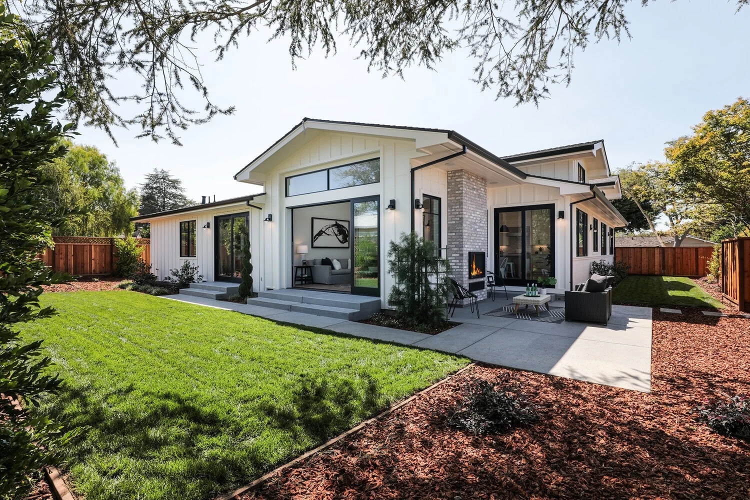 Modern house with large glass windows, outdoor patio with seating, fireplace, well-maintained lawn, and surrounding trees.
