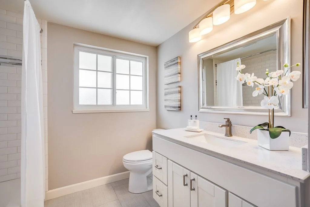 Modern bathroom with white vanity, large mirror, orchid plant, window, and shower curtain.