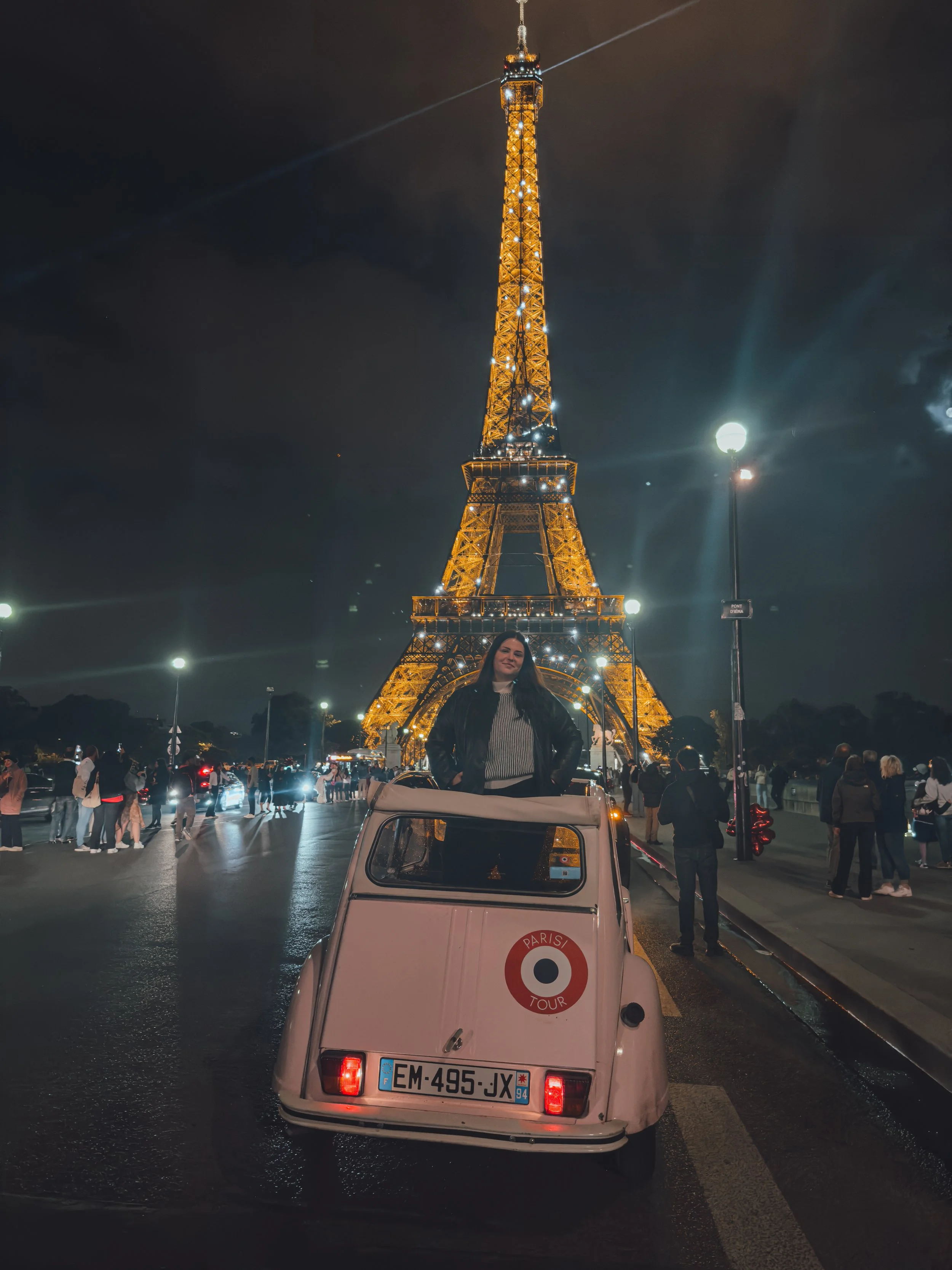 Woman standing in a small white boat with French license plates, parked in front of the Eiffel Tower illuminated at night, with people around and streetlights
