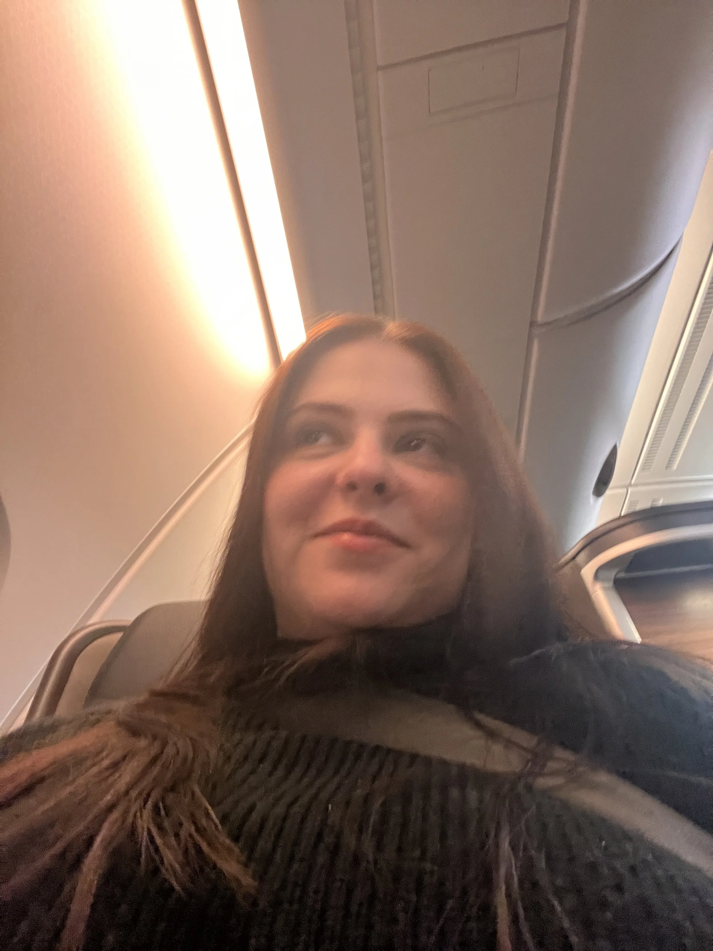 A woman with long brown hair smiling while sitting in an airplane seat, with the overhead compartment and lighting visible above her.