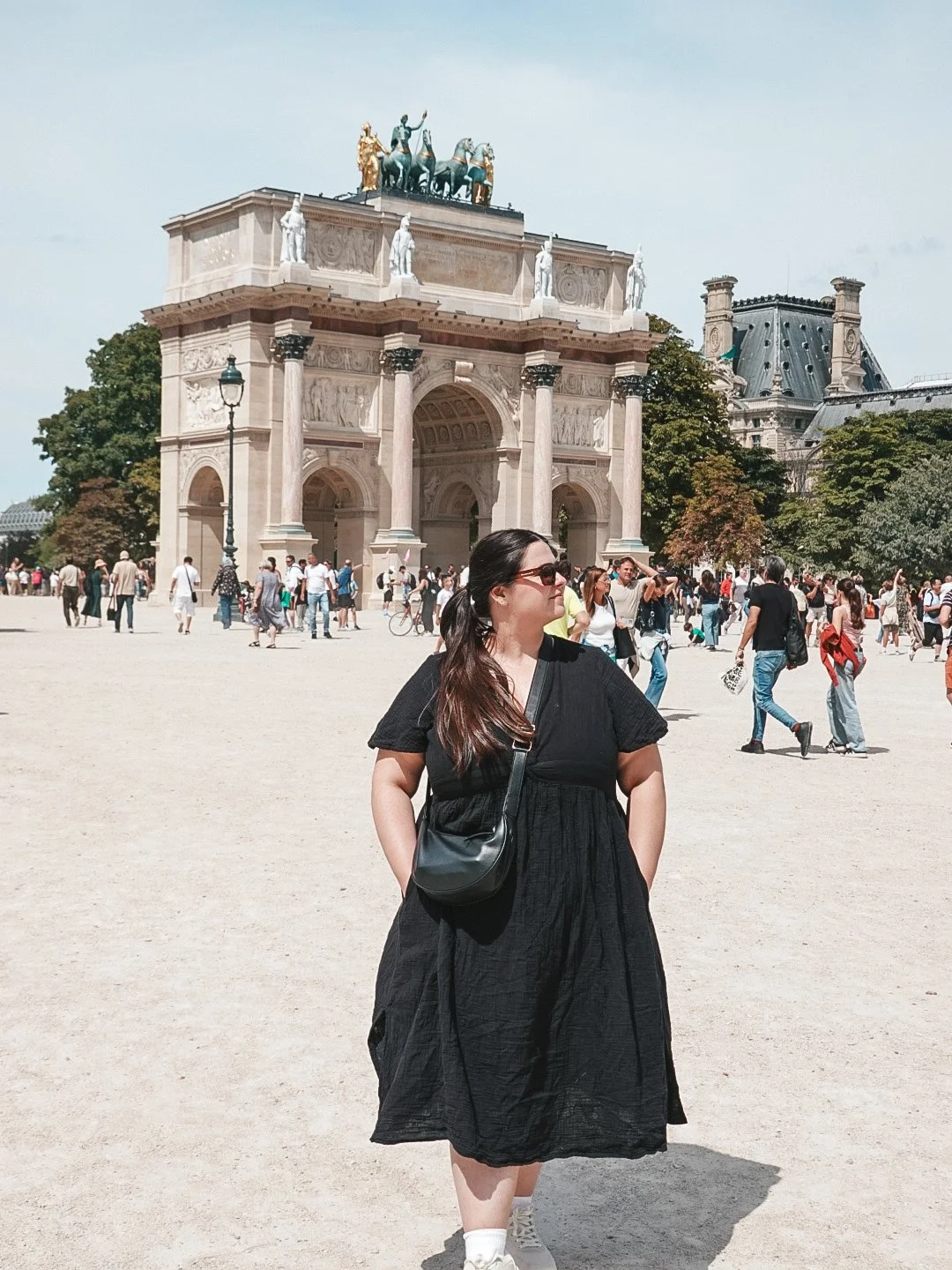 A woman in a black dress and sunglasses stands with her hands in her pockets in front of the Arc de Triomphe du Carrousel in Paris, France, with a crowd of tourists around and trees in the background.