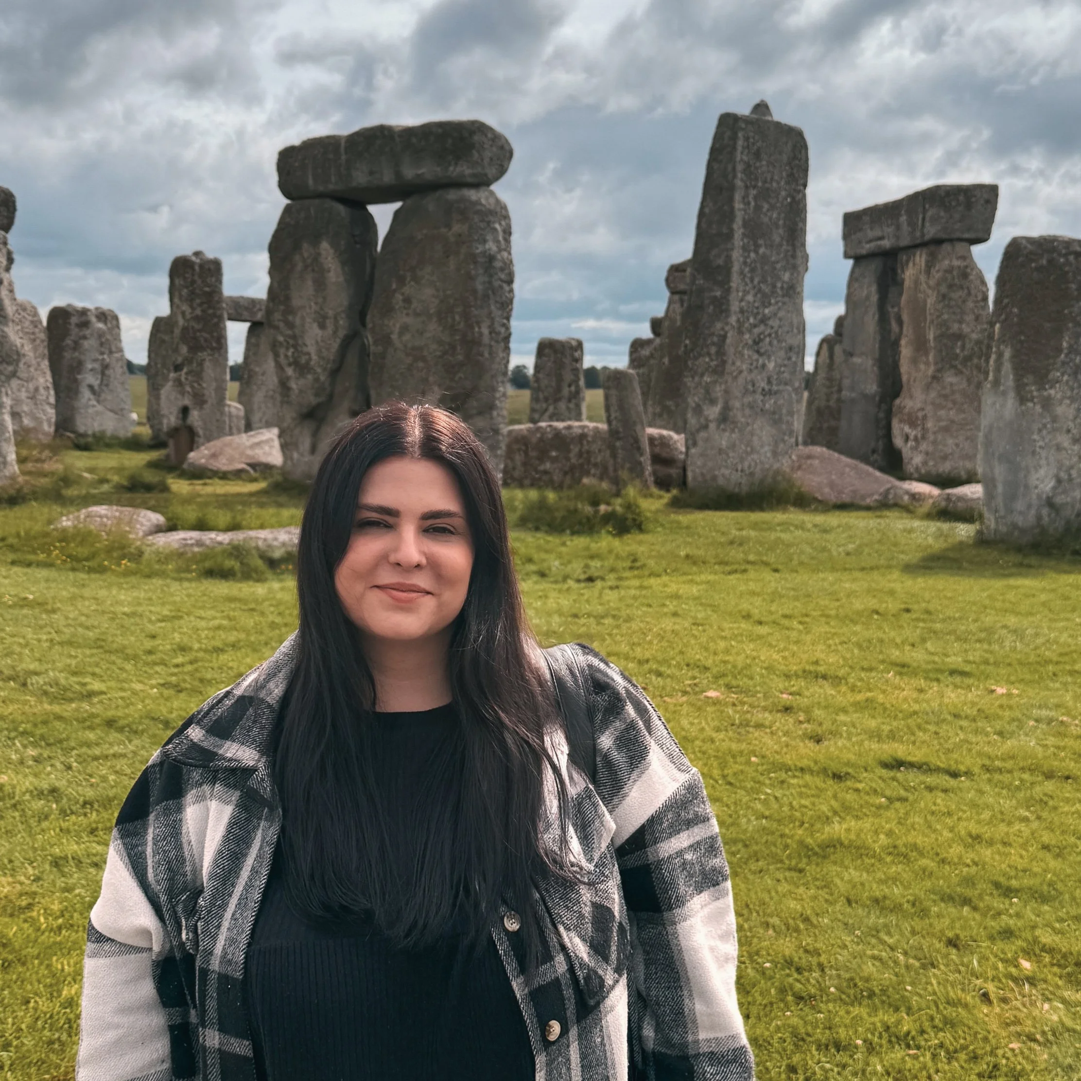 A woman with long dark hair smiling at the camera, standing in front of the ancient Stonehenge monument with large upright stones in the background under a cloudy sky.