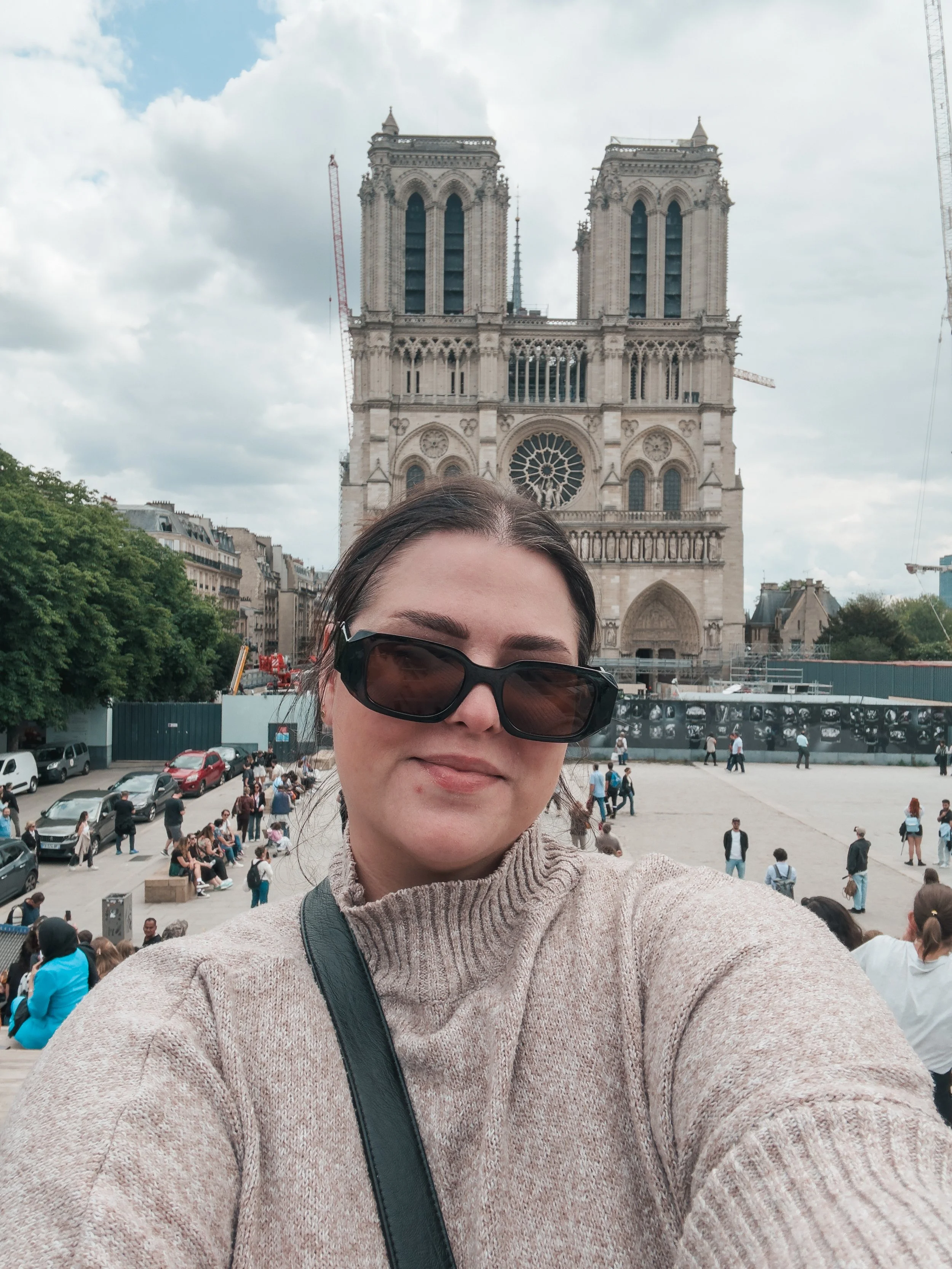 A woman wearing sunglasses and a beige turtleneck sweater taking a selfie in front of Notre Dame Cathedral in Paris, with tourists walking and sitting in the background.