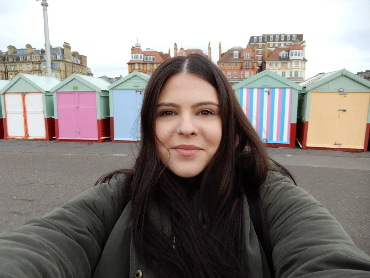 A woman taking a selfie on a beach with colorful beach huts and historic buildings in the background.