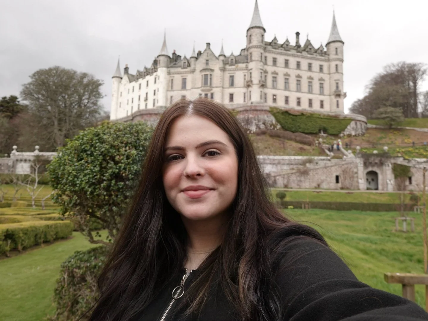 A young woman with long dark hair taking a selfie in front of a castle with green landscape and trees under a cloudy sky.