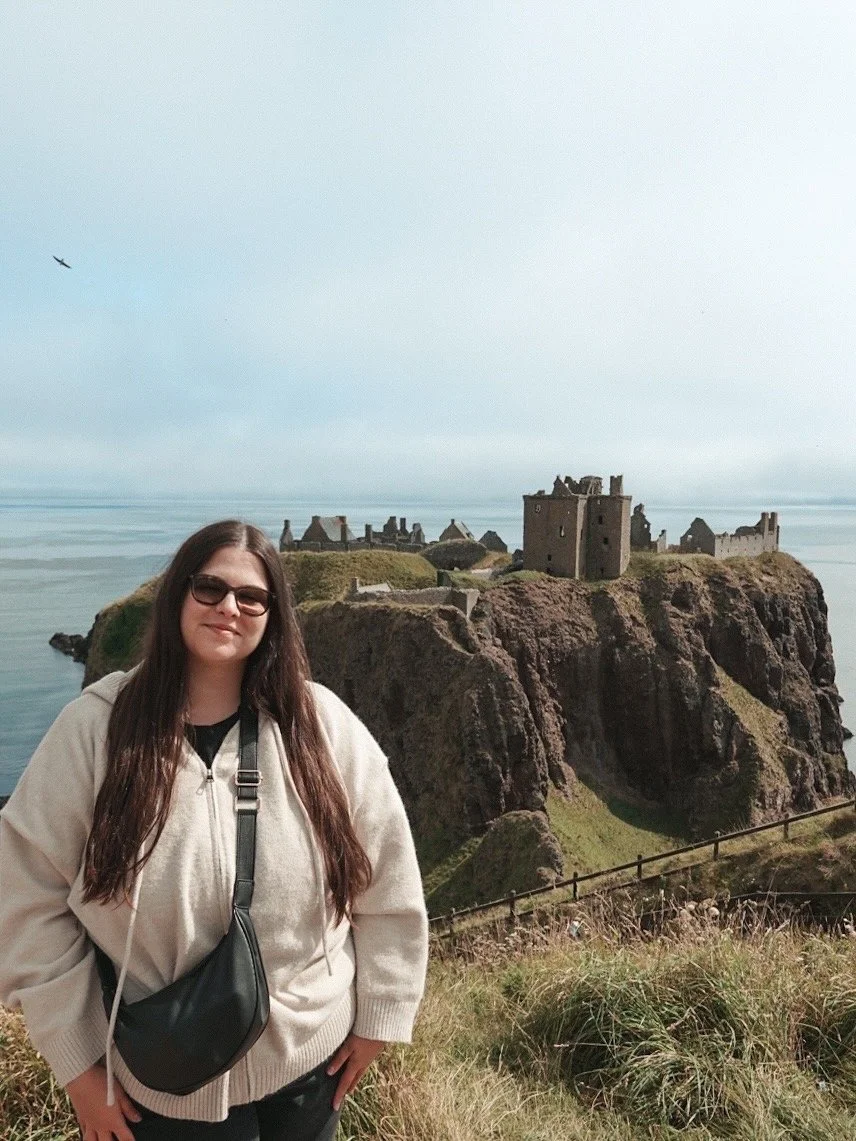 A woman with long dark hair, wearing sunglasses, a beige hoodie, and a black crossbody bag, standing in front of the famous Eilean Donan Castle on a cliff by the water in Scotland.