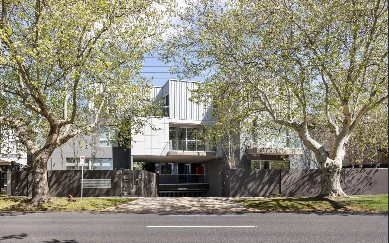 Modern residential building behind a tall dark fence, with two large trees in front casting shadows on the sidewalk, under a clear blue sky.