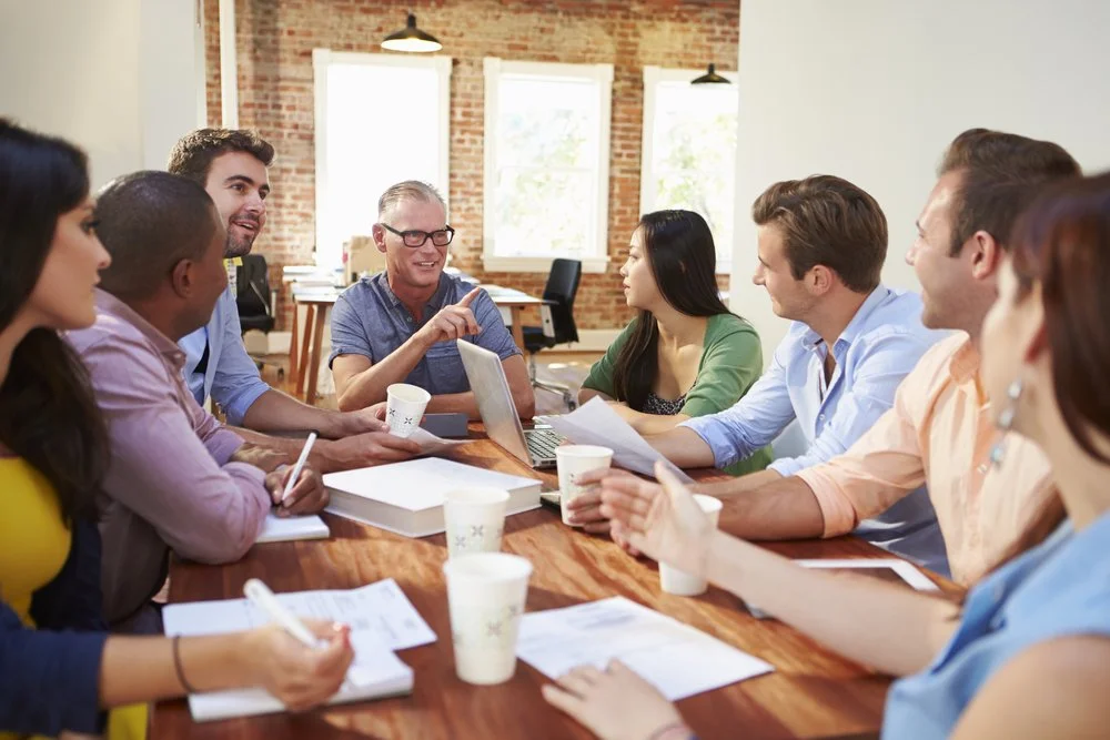 A diverse group of people in a business meeting sitting around a wooden table in an office with exposed brick walls, windows, and bright lighting, engaged in discussion.